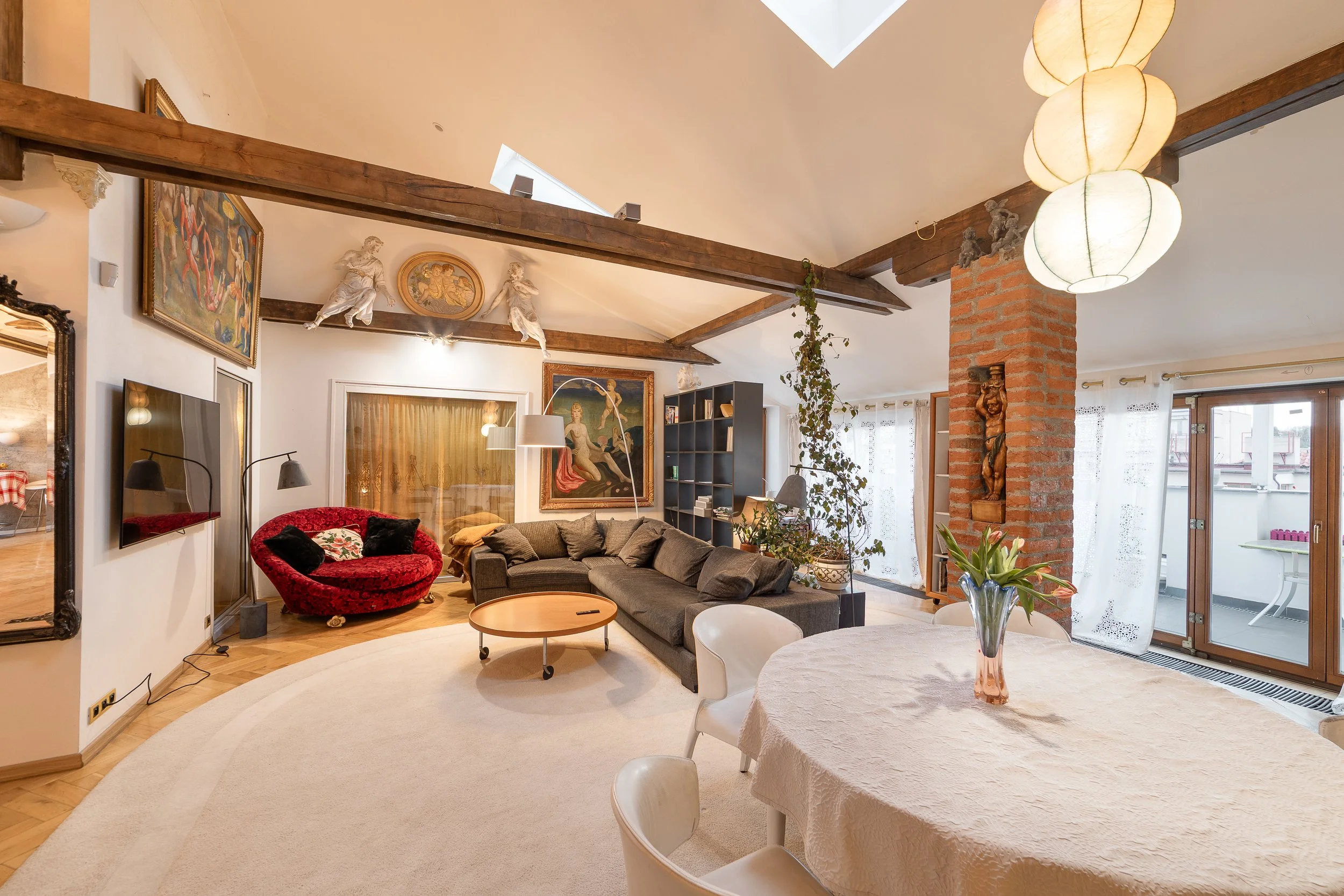 Living room with beige walls, wooden ceiling beams, and skylights. Features include a flat-screen TV, framed artwork, a red velvet armchair, a dark gray sofa, and a round wooden coffee table. A tall bookshelf, houseplants, and a brick column with a c