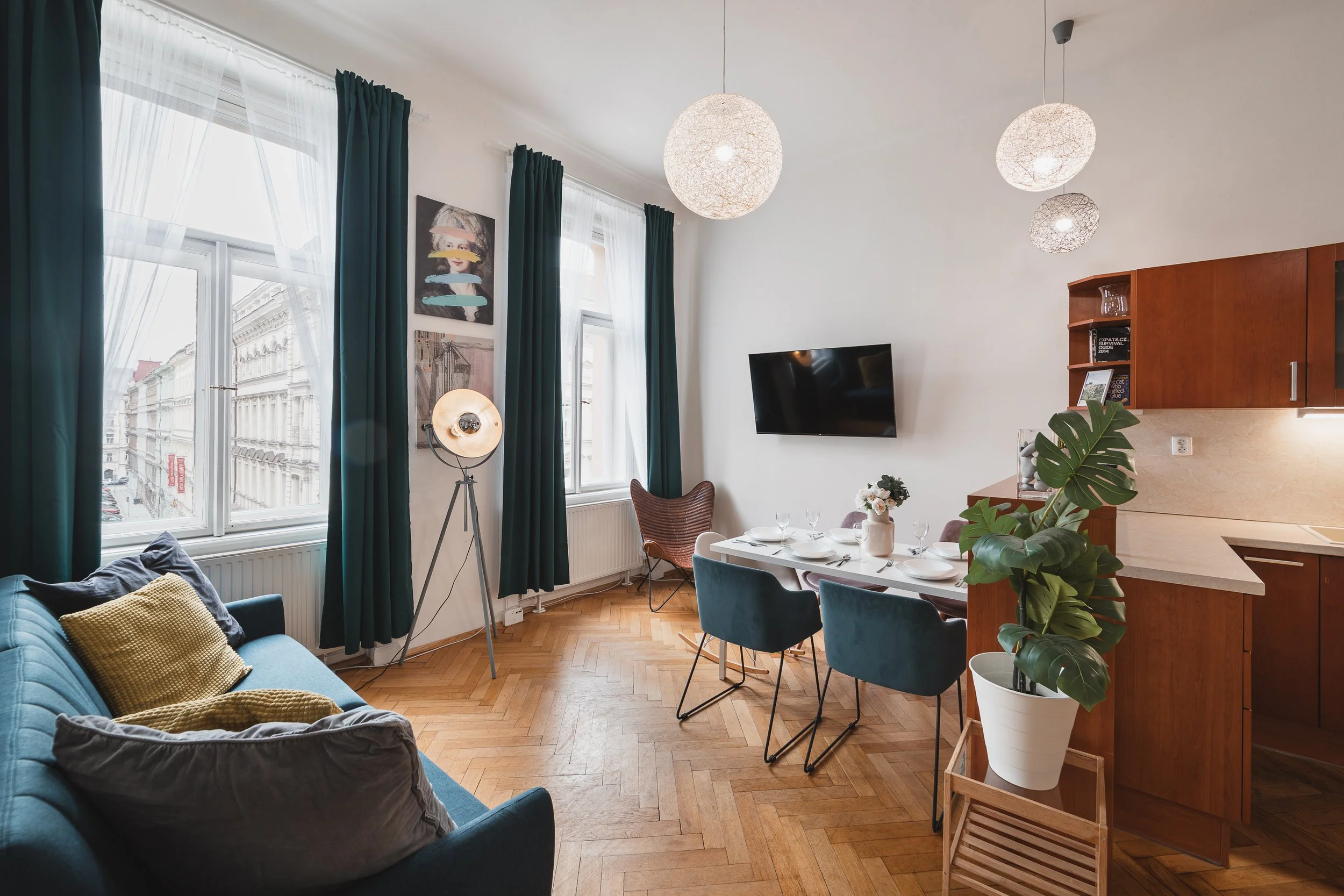 Living room with large windows, dark green curtains, a blue sofa with yellow and gray pillows, a floor lamp with a round shade, a wall-mounted flat screen TV, a wooden cabinet with books and a glass vase, a dining table set with white plates and glas