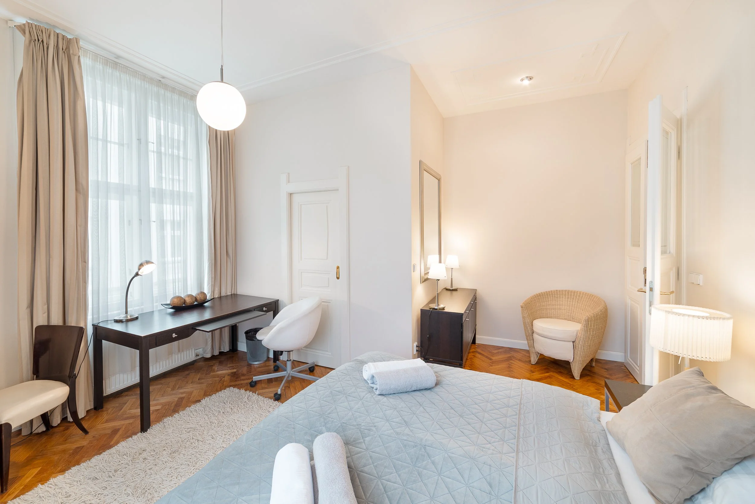 Elegantly decorated bedroom with large window dressed in sheer white curtains and beige drapes, a dark wooden desk, and light-colored walls. The room features a plush bed with light gray bedding, a white upholstered chair, bedside lamps, and wicker a