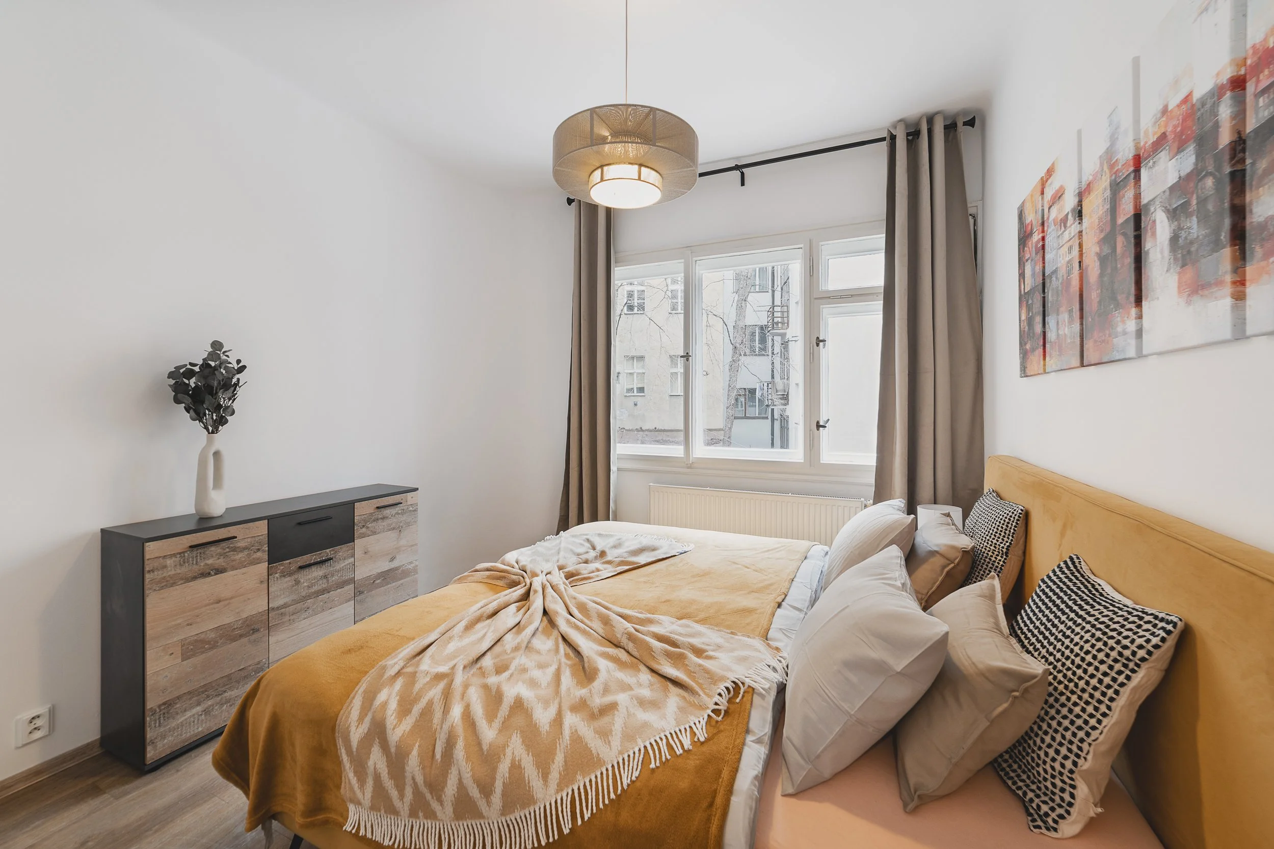 A cozy bedroom with a large window, beige curtains, and modern bedroom decor including a bed with a yellow headboard, plush pillows, and warm-colored blankets. There is a abstract cityscape painting on the wall and a black dresser with decorative ite