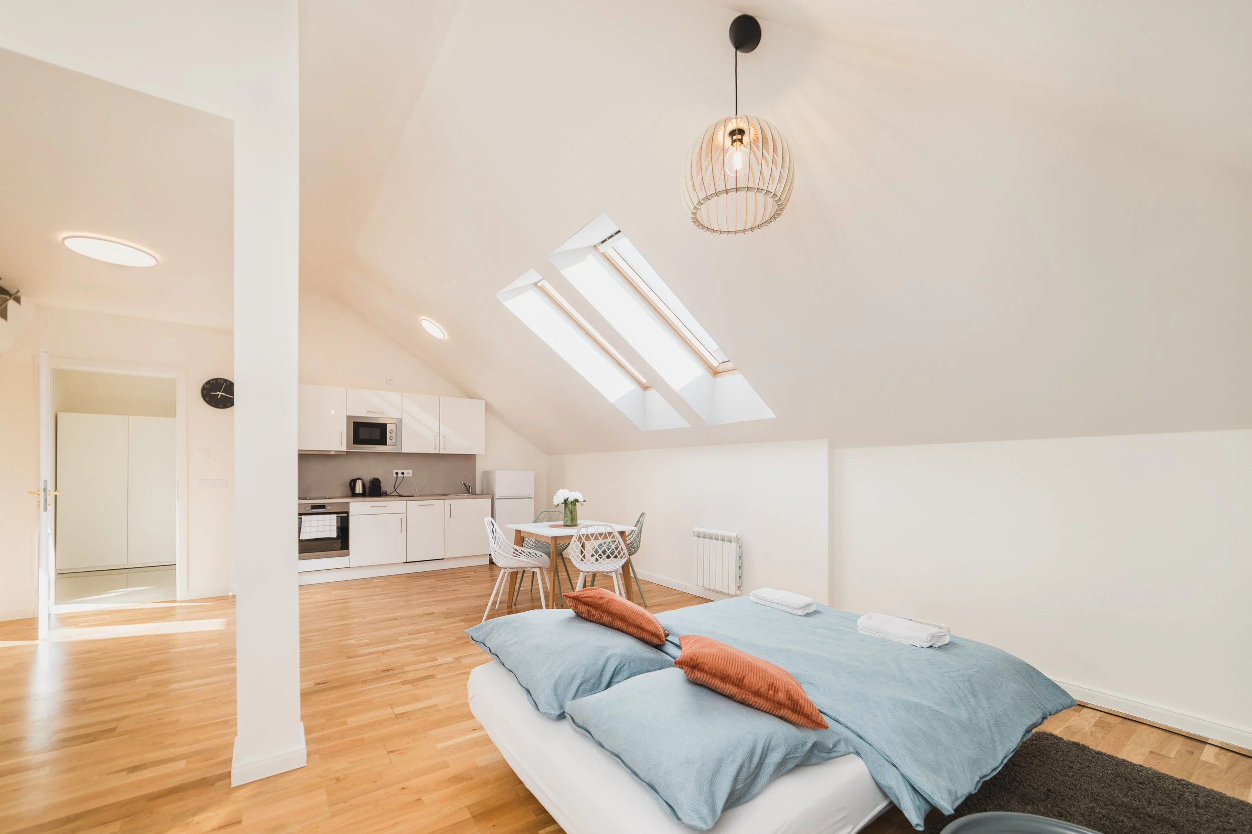 A cozy studio apartment with a slanted ceiling, wooden floors, and a skylight. There is a small dining table with four white chairs, a kitchenette with white cabinets, a microwave, and appliances. The bed in the foreground has blue bedding and orange