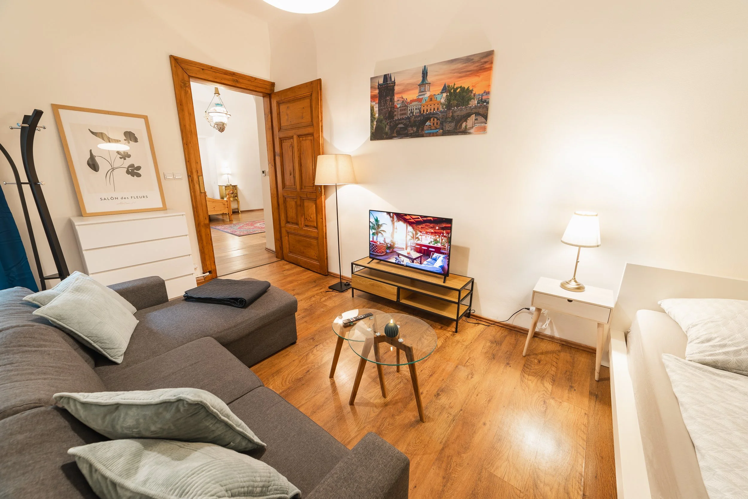 Living room with a gray sofa, white pillows, a round wooden coffee table with decorative items, a TV on a wooden stand, lamps, framed art on the wall, and an open door leading to a bedroom with additional lamp lighting.