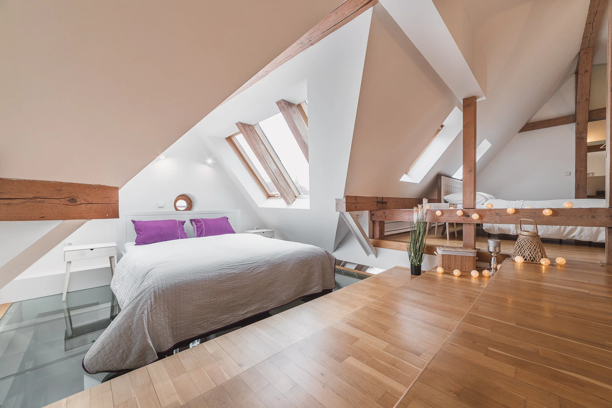 A bedroom with a sloped ceiling, skylight windows, white walls, and wooden beams. It features a bed with white bedding and purple pillows, white bedside tables, and decorative string lights on a wooden surface. There is a small round window and vario