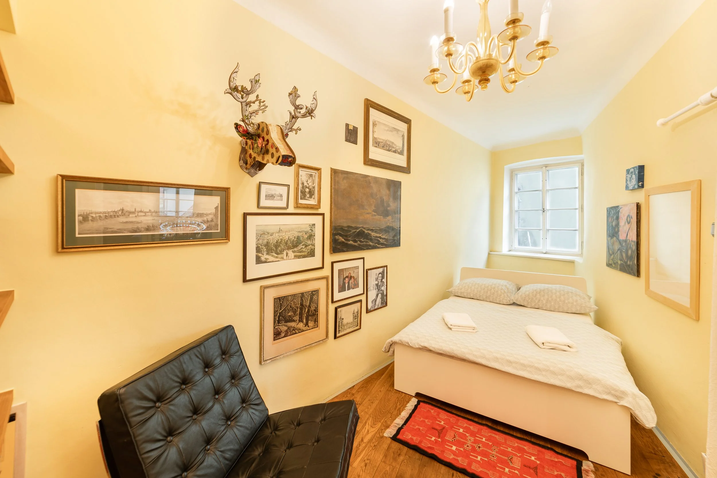 A cozy bedroom with yellow walls decorated with framed artwork, a window letting in natural light, a bed with a white bedspread, a black leather tufted sofa, and a red decorative rug on a wooden floor. There is also a chandelier hanging from the ceil