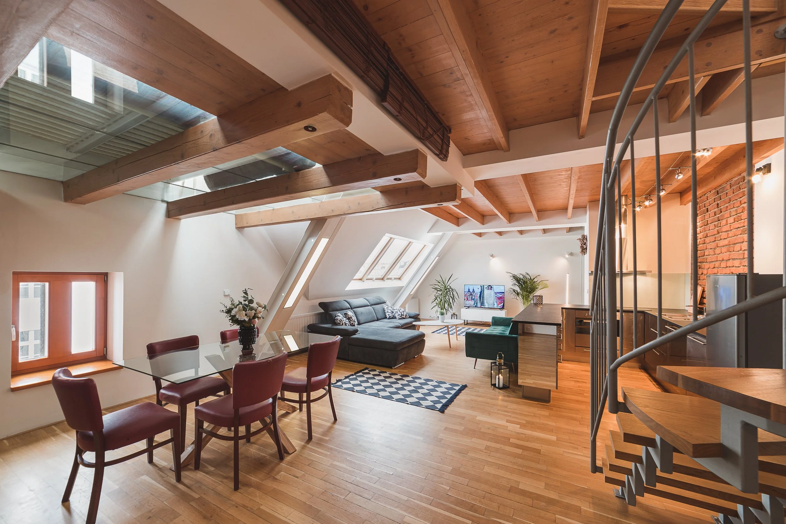 Interior view of a modern living room and dining area with wooden floors, vaulted wooden ceiling, brick wall, large windows, and contemporary furniture including a gray sofa, a glass dining table with pink chairs, and potted plants.