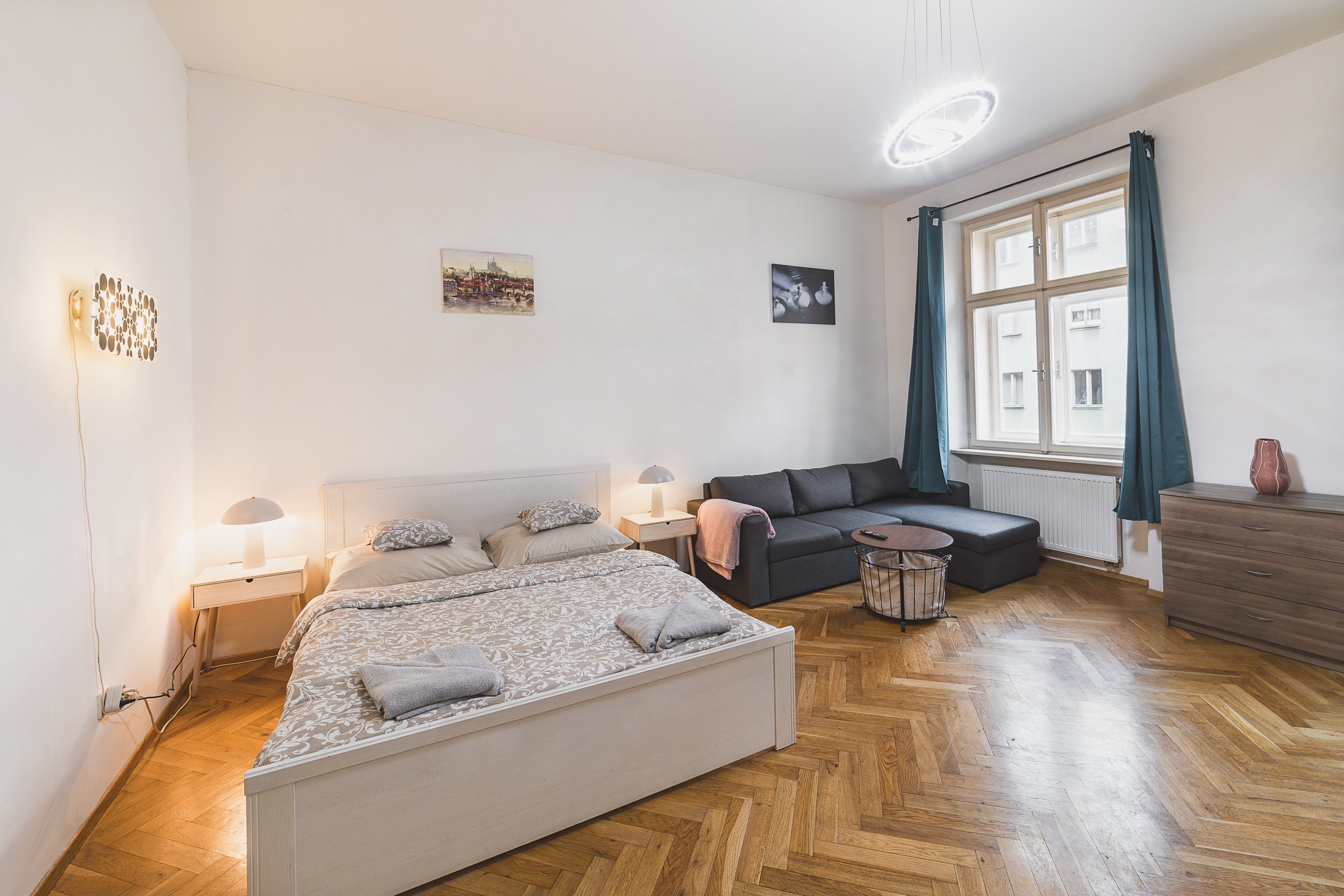 A furnished apartment living room with a bed, side tables, sofa, armchair, window with blue curtains, and wall art.
