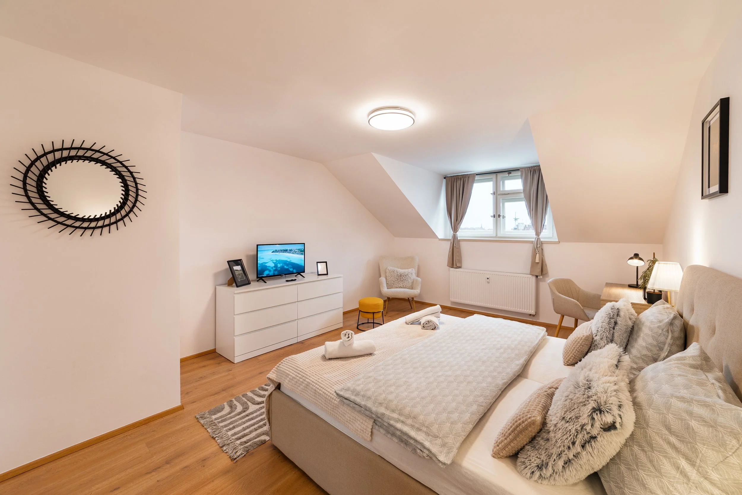 A cozy bedroom with a sloped ceiling featuring a wall-mounted black decorative mirror, a flat-screen TV on a white dresser, a beige armchair, a window with beige curtains, a bed with rolled towels, fluffy pillows, and a gray and white rug.