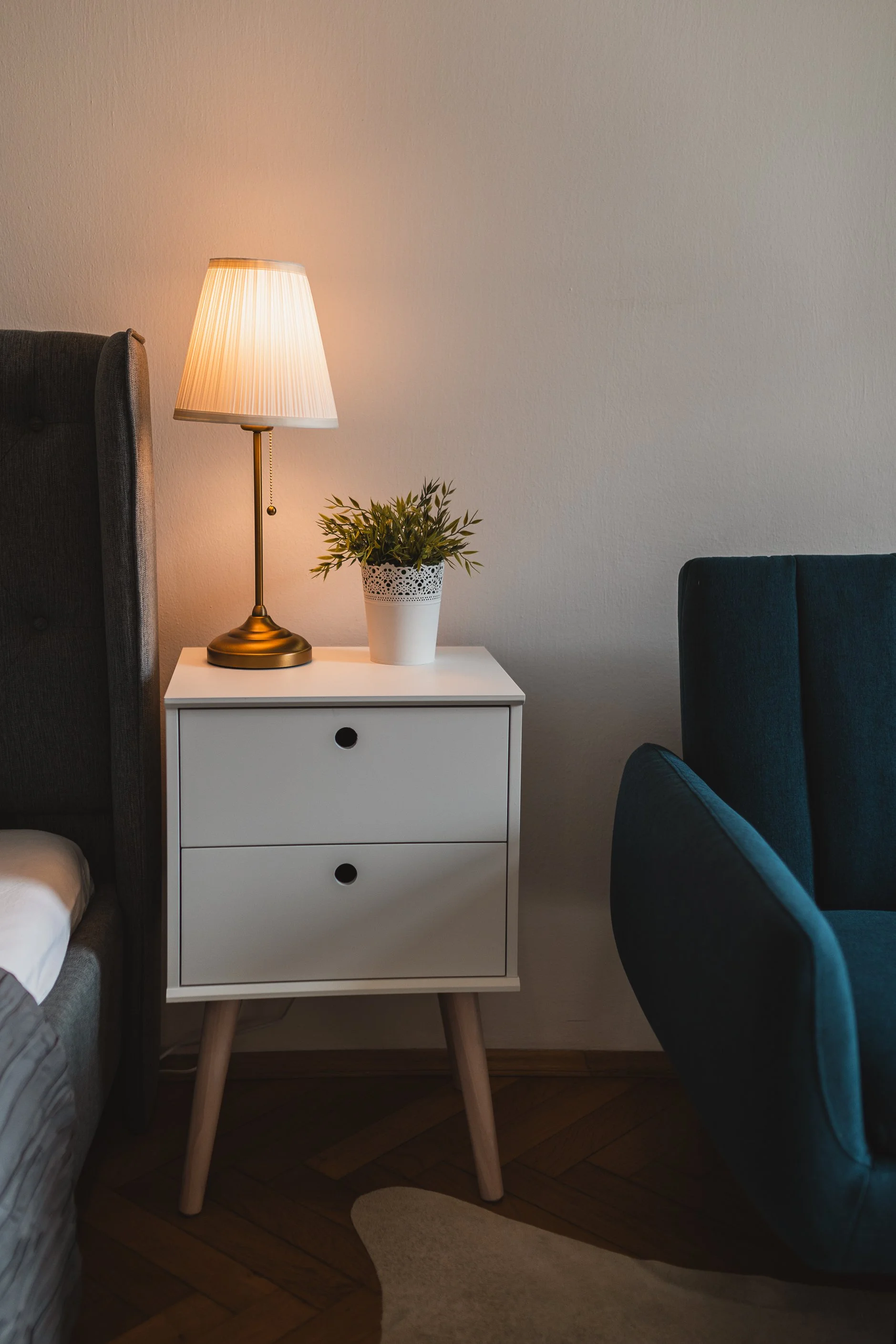 A white nightstand with three drawers and wooden legs, placed next to a bed with a gray headboard, a brass lamp with a pleated lampshade, and a potted green plant, in a room with wooden flooring, beige walls, and a blue upholstered armchair to the ri