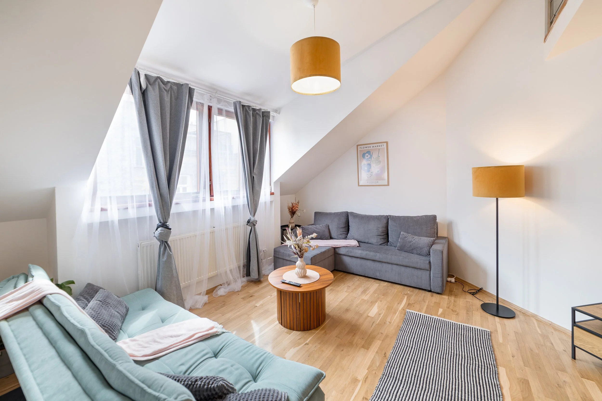 Living room with sloped ceiling, grey sofa, light blue sofa, round wooden coffee table, yellow standing lamp, window with grey curtains, framed artwork on wall, small side table, striped rug, wooden floor.