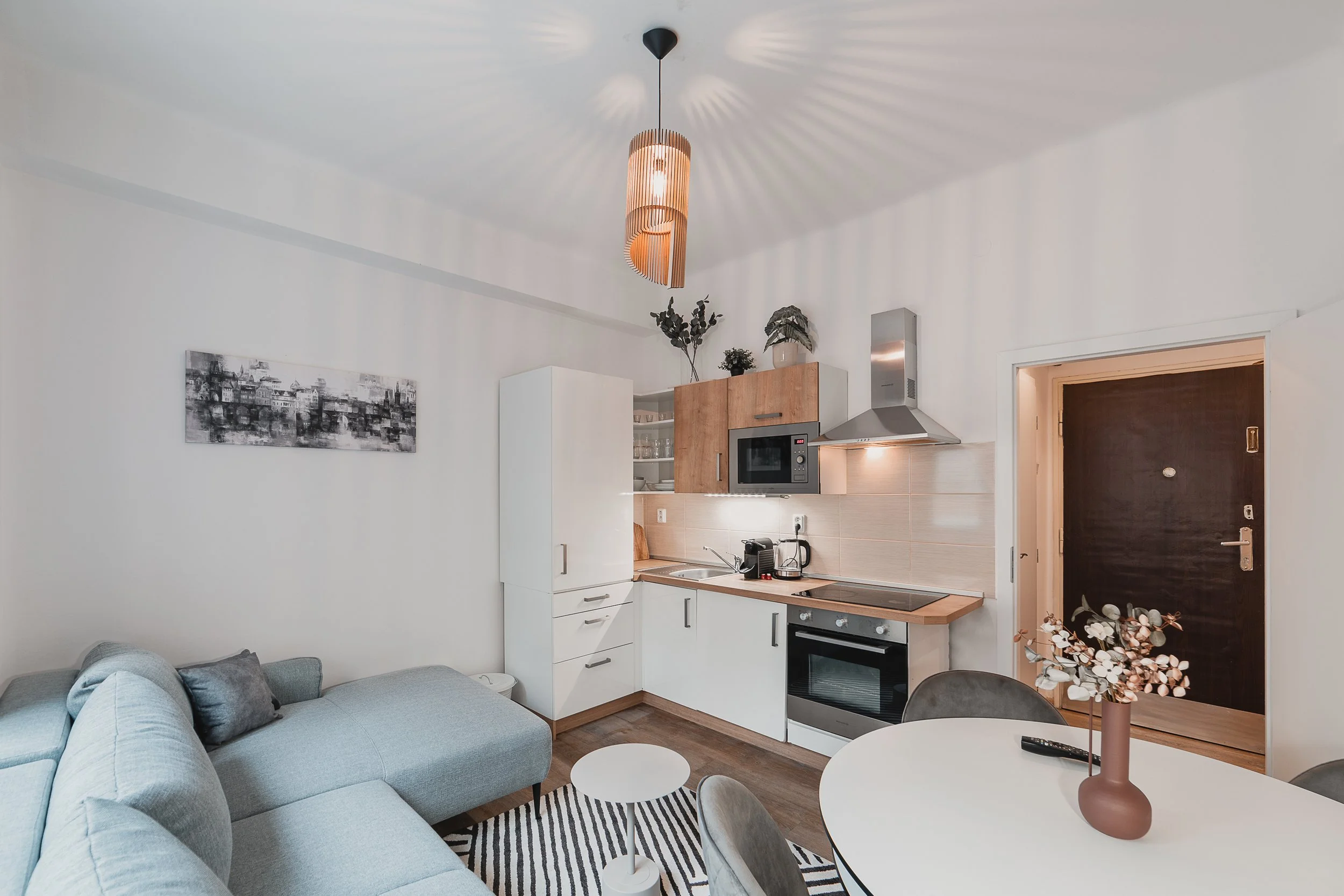 Modern kitchen with white cabinets, wooden accents, and stainless steel appliances, adjacent to a living area with a gray sofa and round white coffee tables, decorated with a pink vase and flowers.