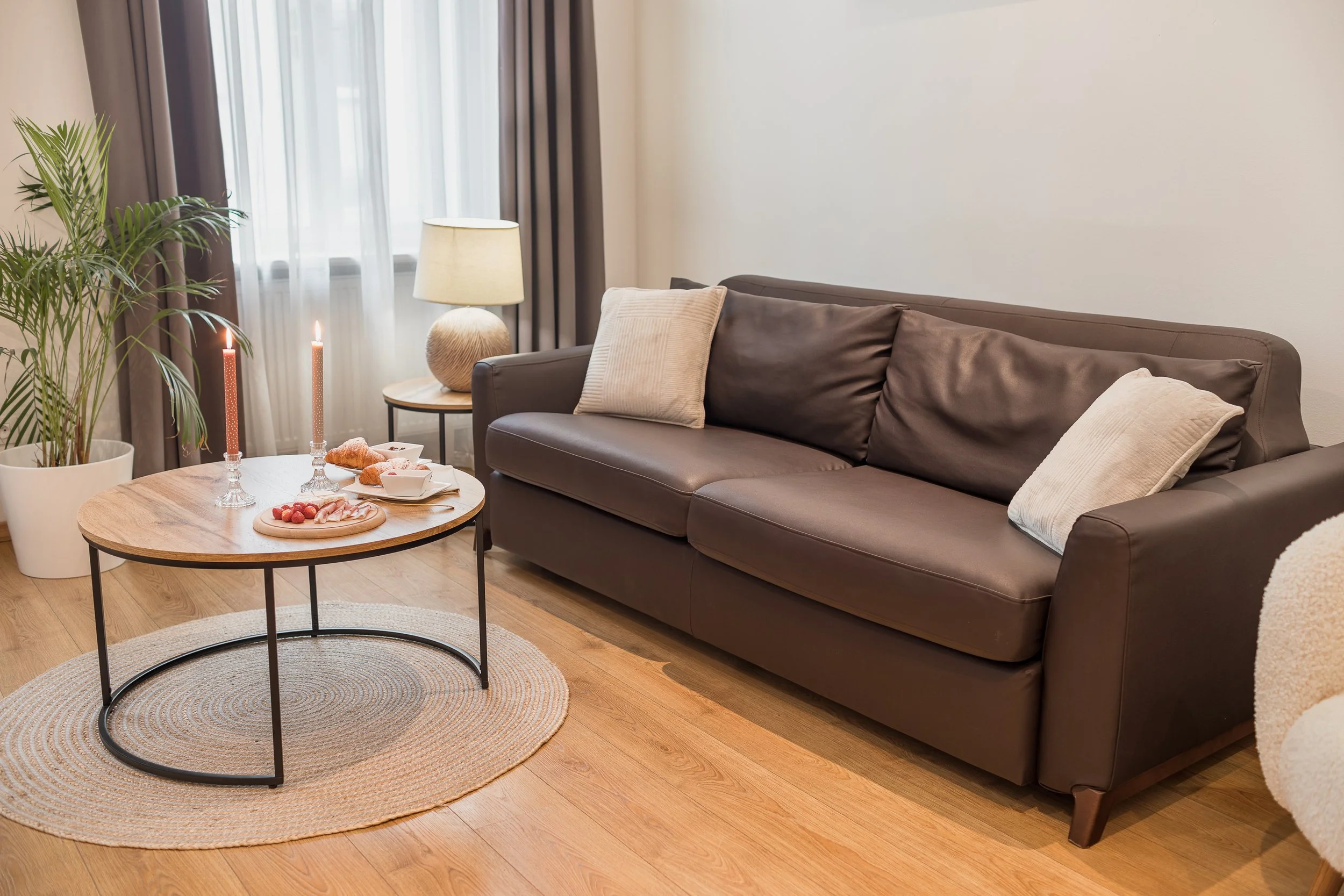 Living room with a brown leather sofa, beige and brown pillows, a braided area rug, a wooden coffee table with snacks, a potted plant, a side table with a lamp, and candles near a window with curtains.