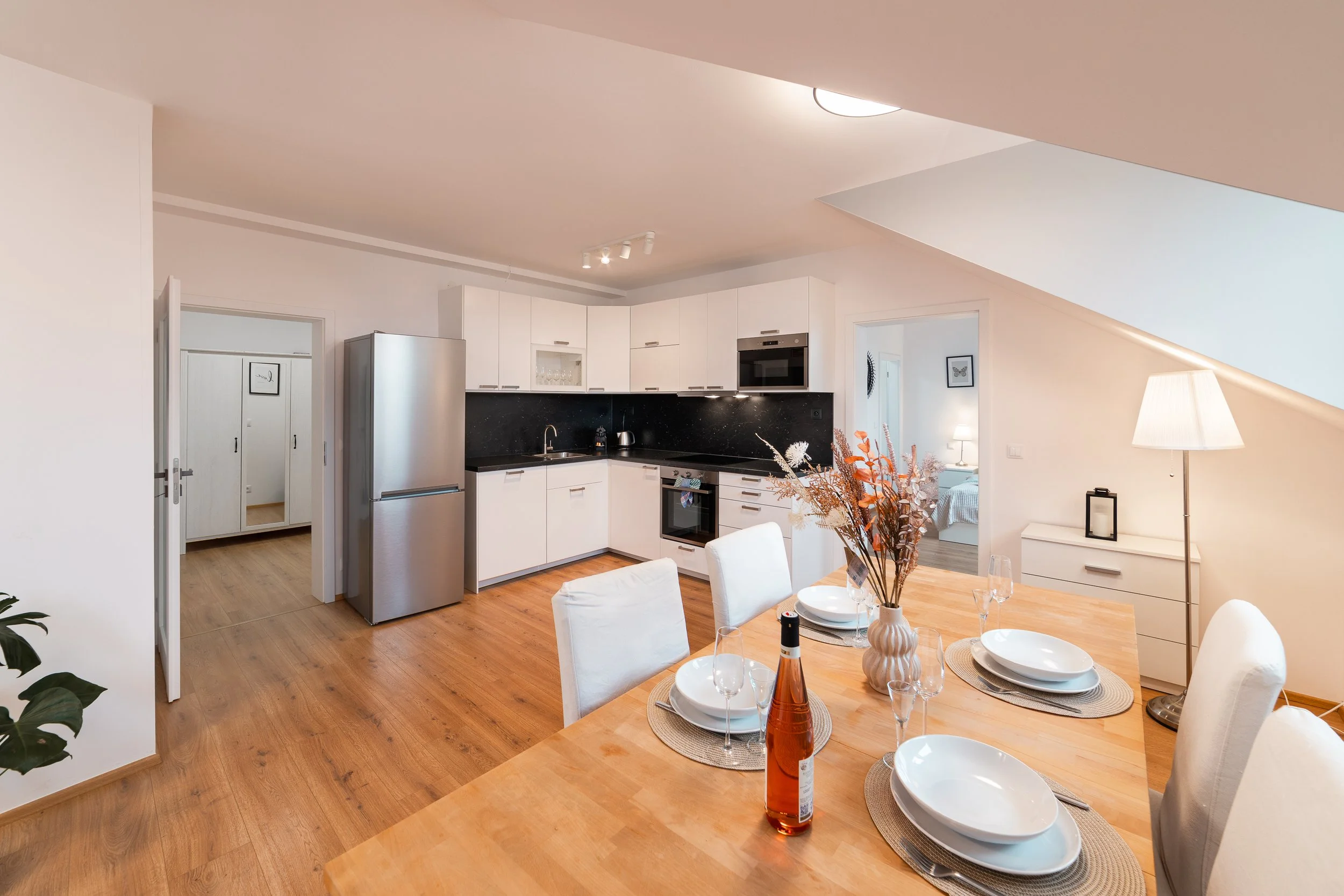 Modern open-concept kitchen and dining area with wooden floors, white cabinets, stainless steel appliances, and a dining table set with white plates, wine glasses, and a decorative orange vase with flowers.