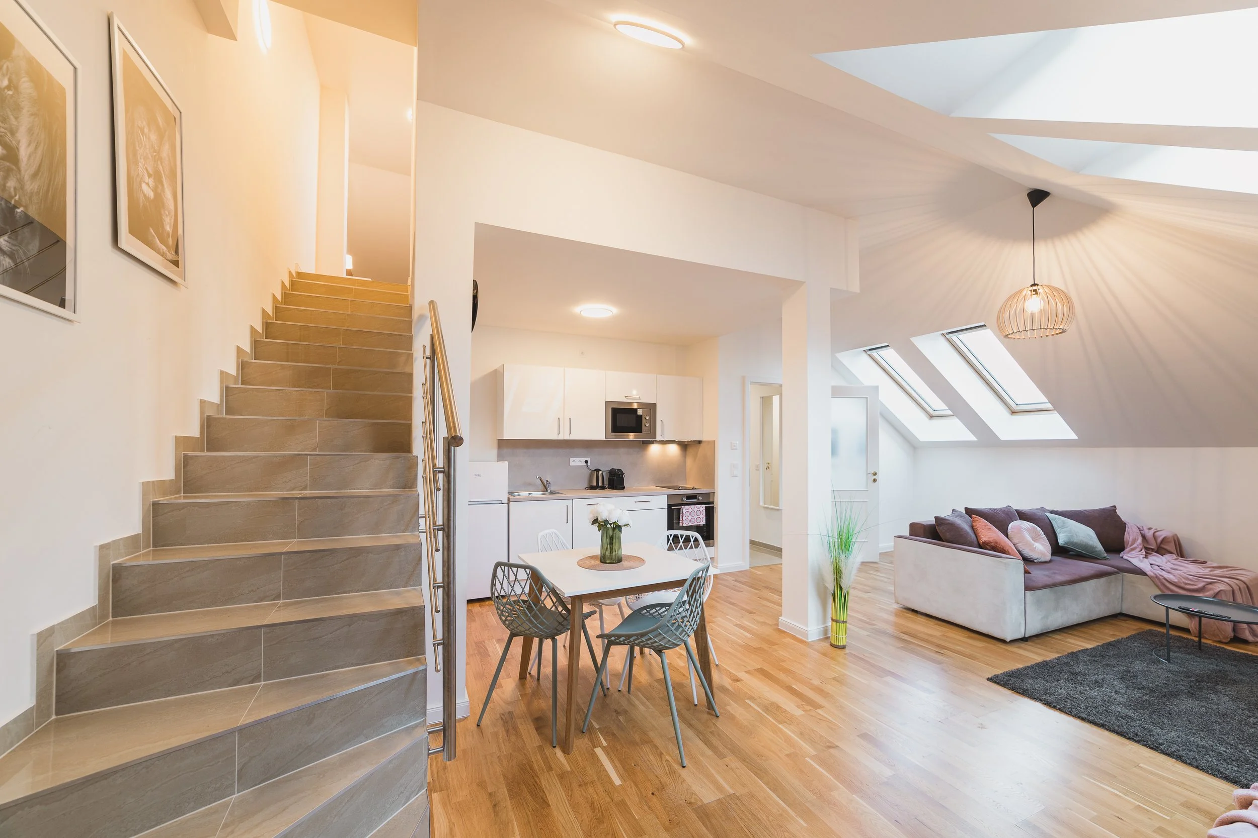 Bright open-plan living space with a staircase, small dining table, kitchen area, and a seating area with a sofa near skylights and modern lighting fixtures.