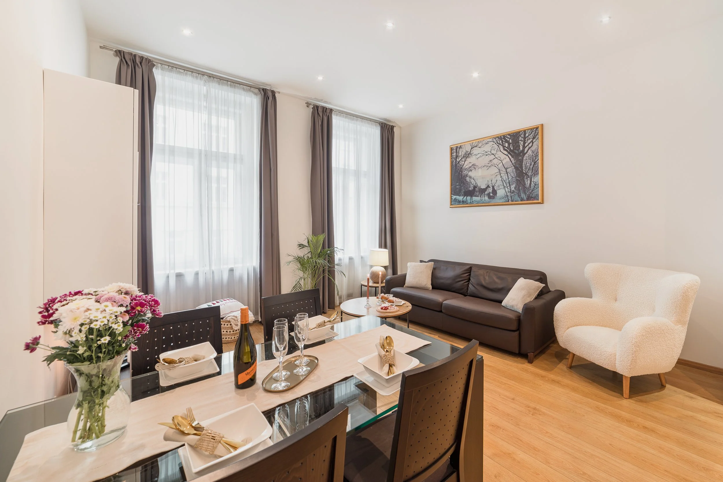 Living room with windows, curtains, a painting, a sofa, a table with flowers, wine bottles, and dining setup.