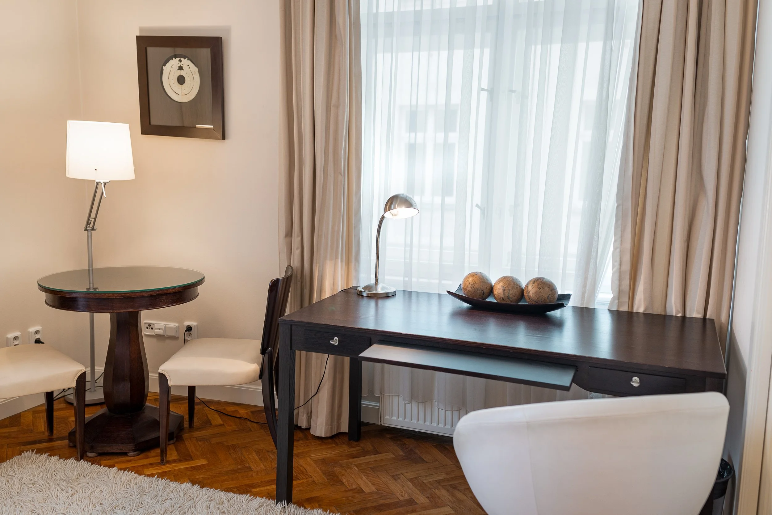 Interior of a living space with a window covered by sheer and curtain drapes, a dark wooden table with a small lamp, a decorative tray with spherical objects, a framed artwork on the wall, and chairs nearby, with hardwood flooring and a textured whit