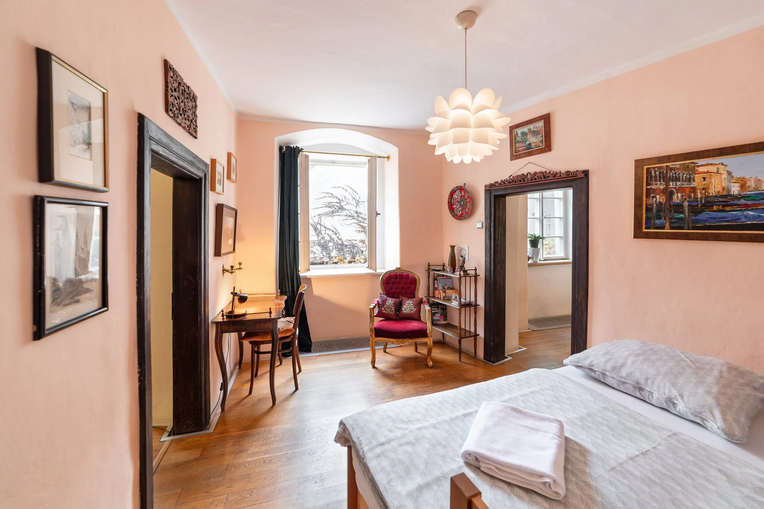 A cozy bedroom with peach-colored walls, a bed with white bedding, and a wooden floor. There are framed pictures on the walls, a window with dark curtains, and a chandelier hanging from the ceiling. The room has antique furniture, including a red vel