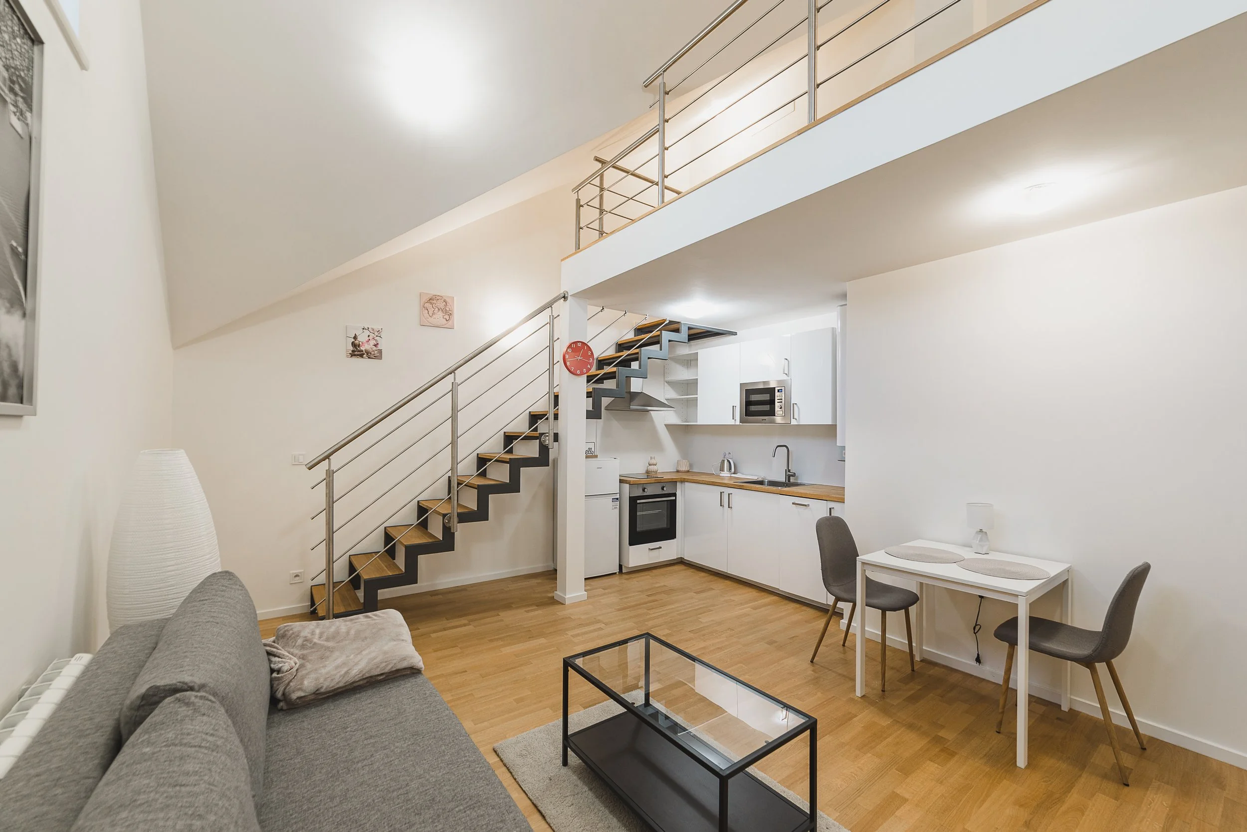 Interior view of a modern apartment with an open kitchen, staircase, and dining area. The space features white walls, wooden flooring, and minimalist decor.