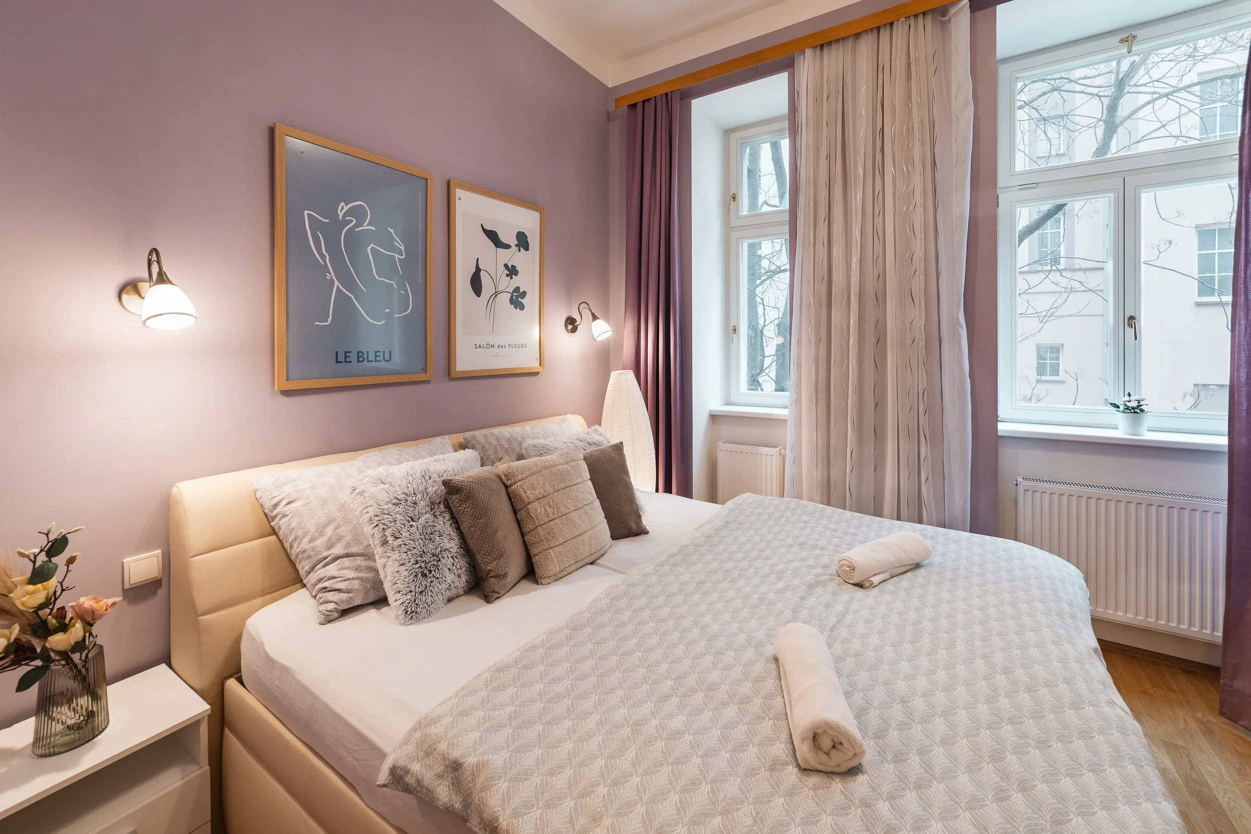 A cozy bedroom with a beige upholstered bed covered in light-colored bedding and multiple decorative pillows. There are two rolled towels at the foot of the bed. A white nightstand with a floral arrangement in a glass vase is beside the bed. The wall