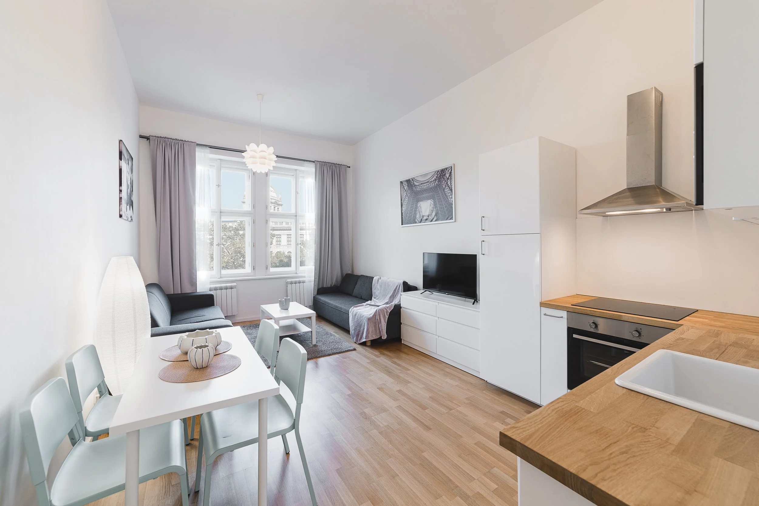 A living room with a dining table set for four, a sofa, a television, and a modern kitchen area with white cabinets and a stove near a window with curtains.