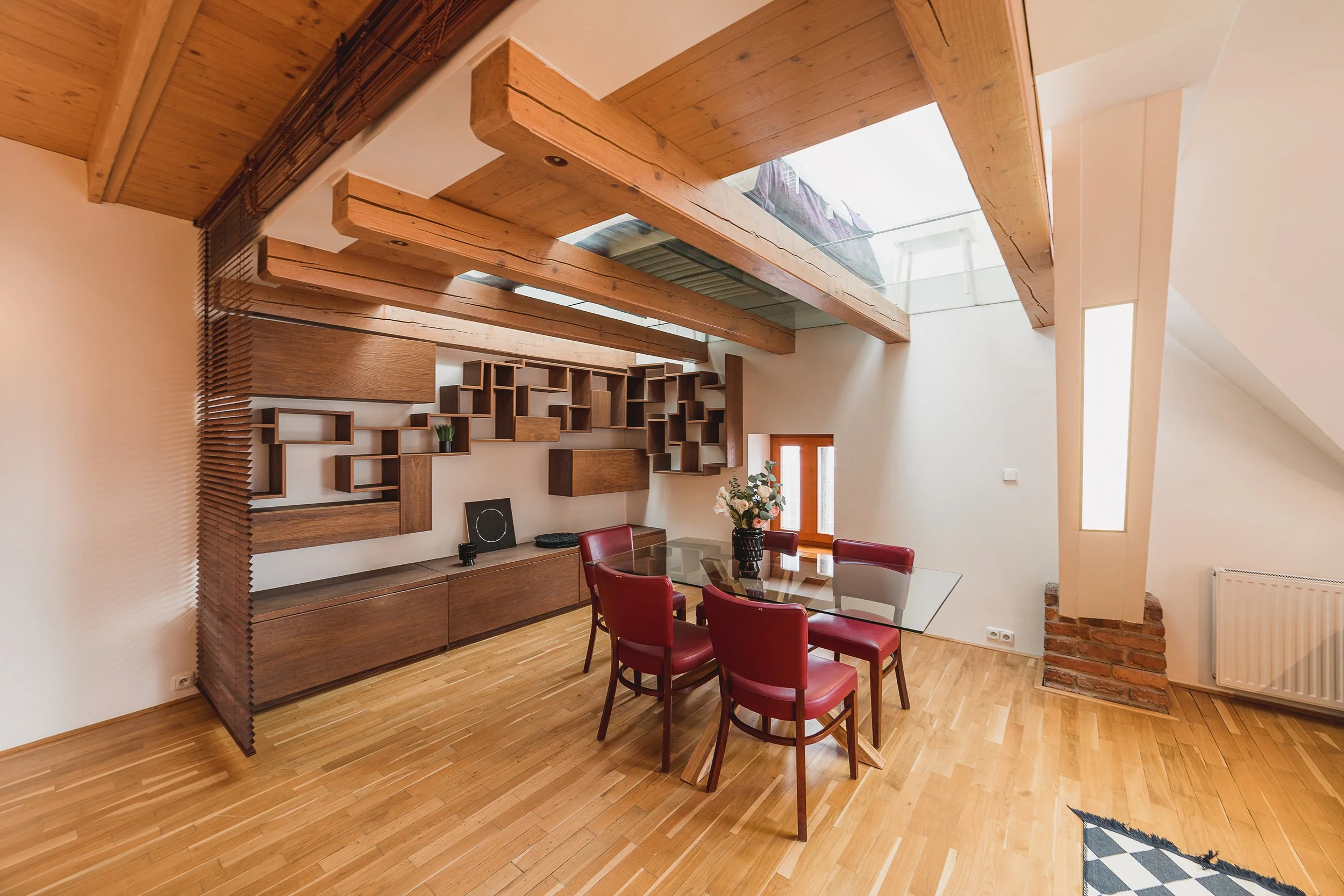 A cozy dining room with a wooden floor and exposed brick wall. Features a large glass table with red upholstered chairs, wall-mounted wooden shelves, and a skylight ceiling letting in natural light.