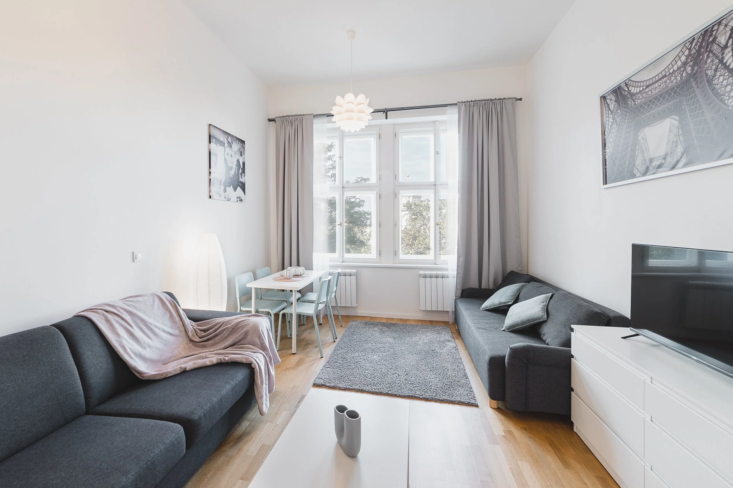 Living room with white walls, large windows with curtains, two gray sofas, a small white dining table with light blue chairs, a TV on a white stand, and wall art, with hardwood floor and a gray rug.