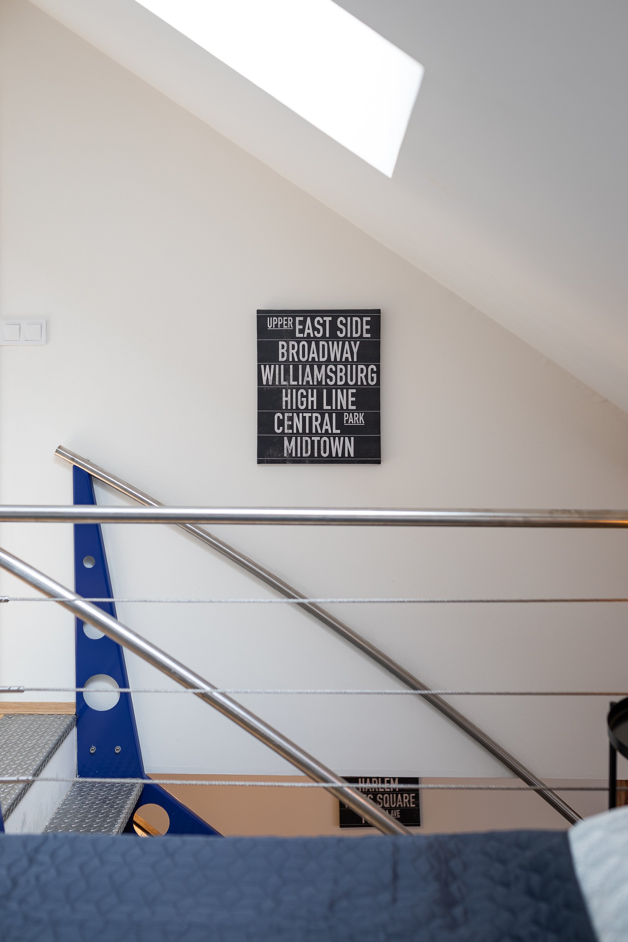 Interior view of a stairway with a black and white signage displaying names of New York City neighborhoods, including East Side, Broadway, Williamsburg, High Line, Central Park, and Midtown, along with part of a grey handrail and blue support structu