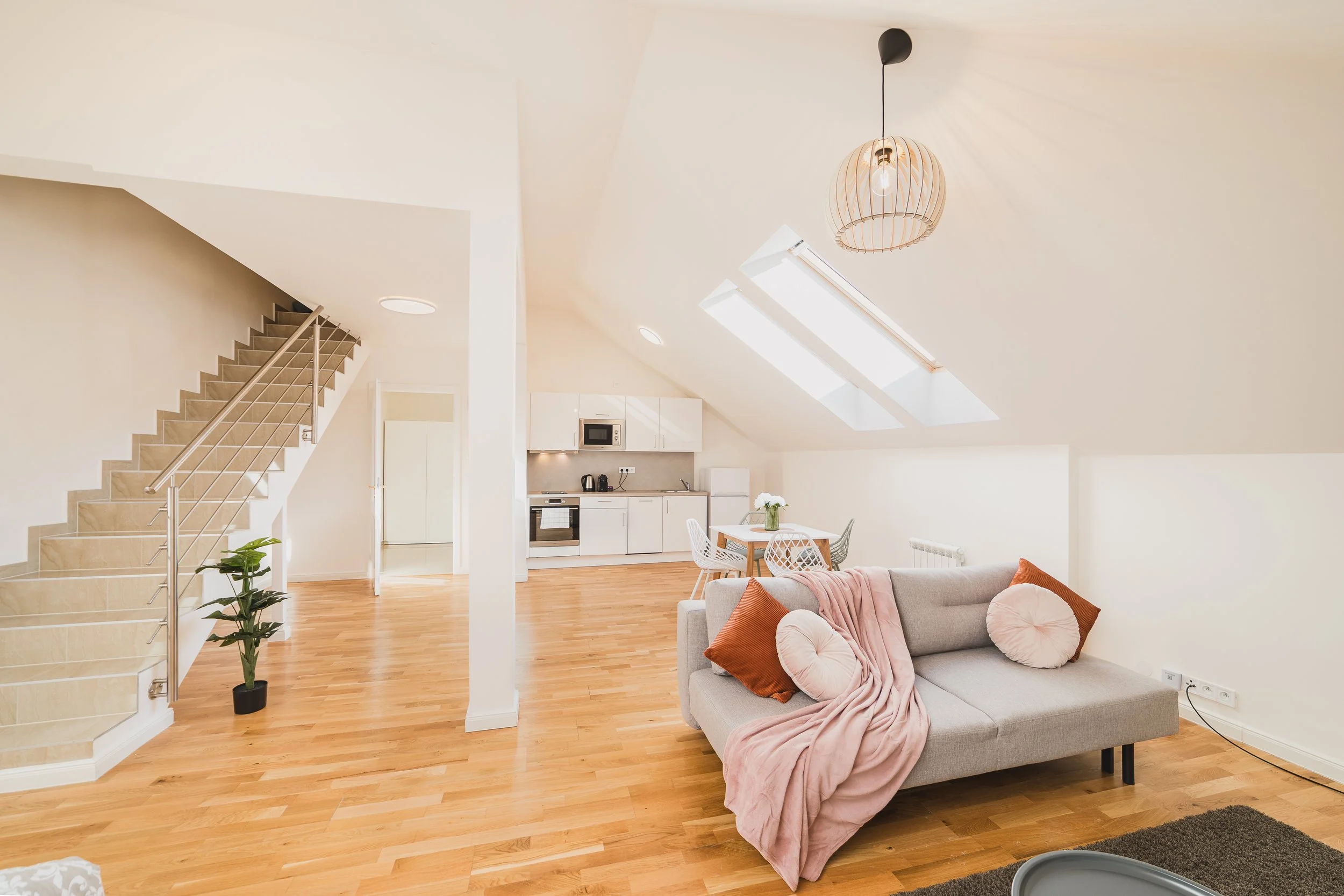 Open-concept living area with beige sofa, pink blanket, and cushions, adjacent to a dining space and kitchen with white cabinets, microwave, and small appliances, under skylights and pendant light.