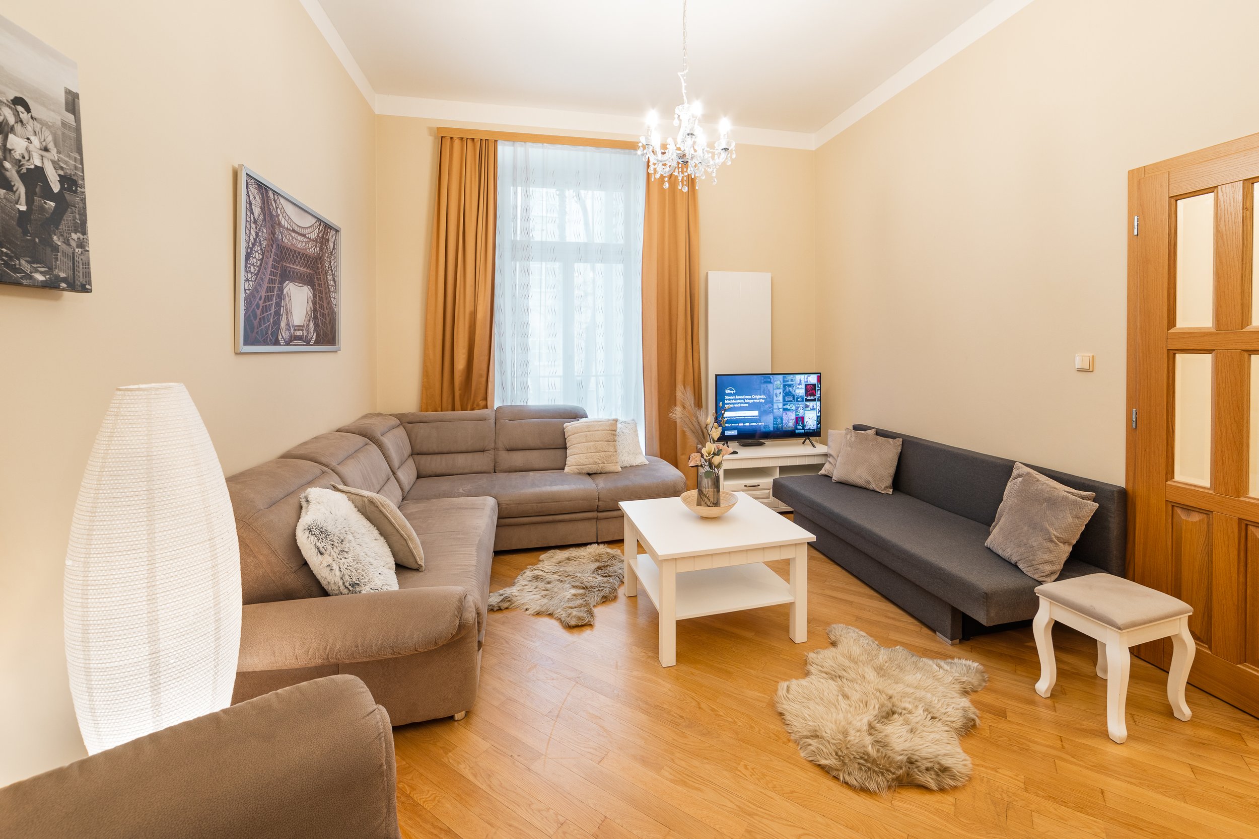 Living room with beige walls, large window with white sheer curtains and orange drapes, wooden floor, two beige sofas, one dark grey sofa, white coffee table, television, framed photos, chandelier, lamp, decorative pillows, and small fur rugs.
