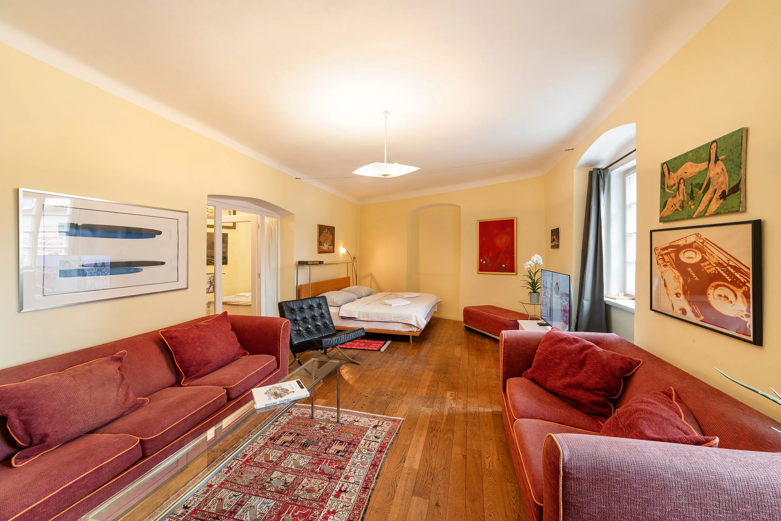 A living room with yellow walls, red sofas, framed artwork, a television, a bed in the background, and windows with curtains.