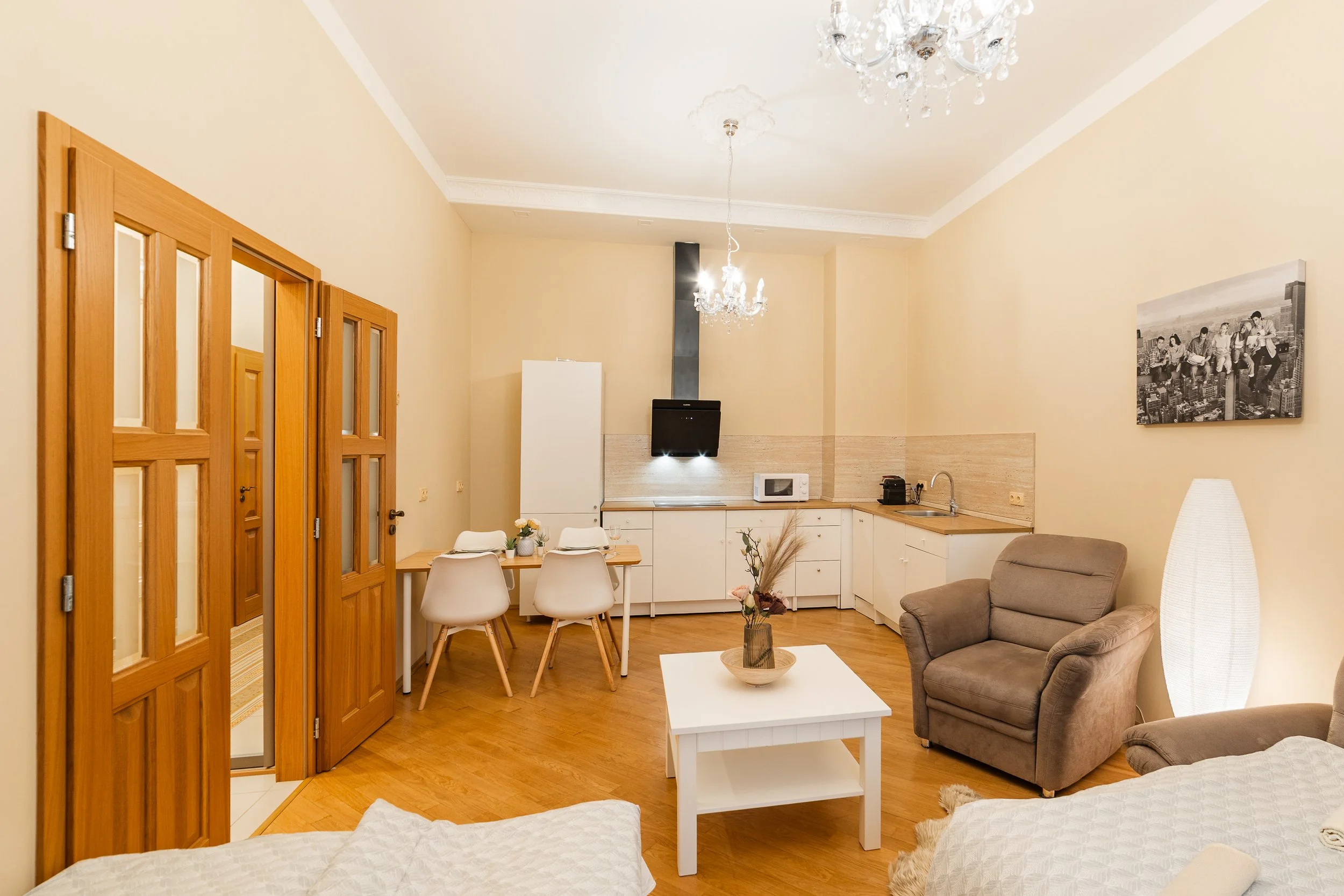 A cozy living room with cream walls, wooden accents, a chandelier, a sofa, armchair, coffee table with a vase, a dining area with chairs, wall art, kitchen appliances, and hardwood flooring.