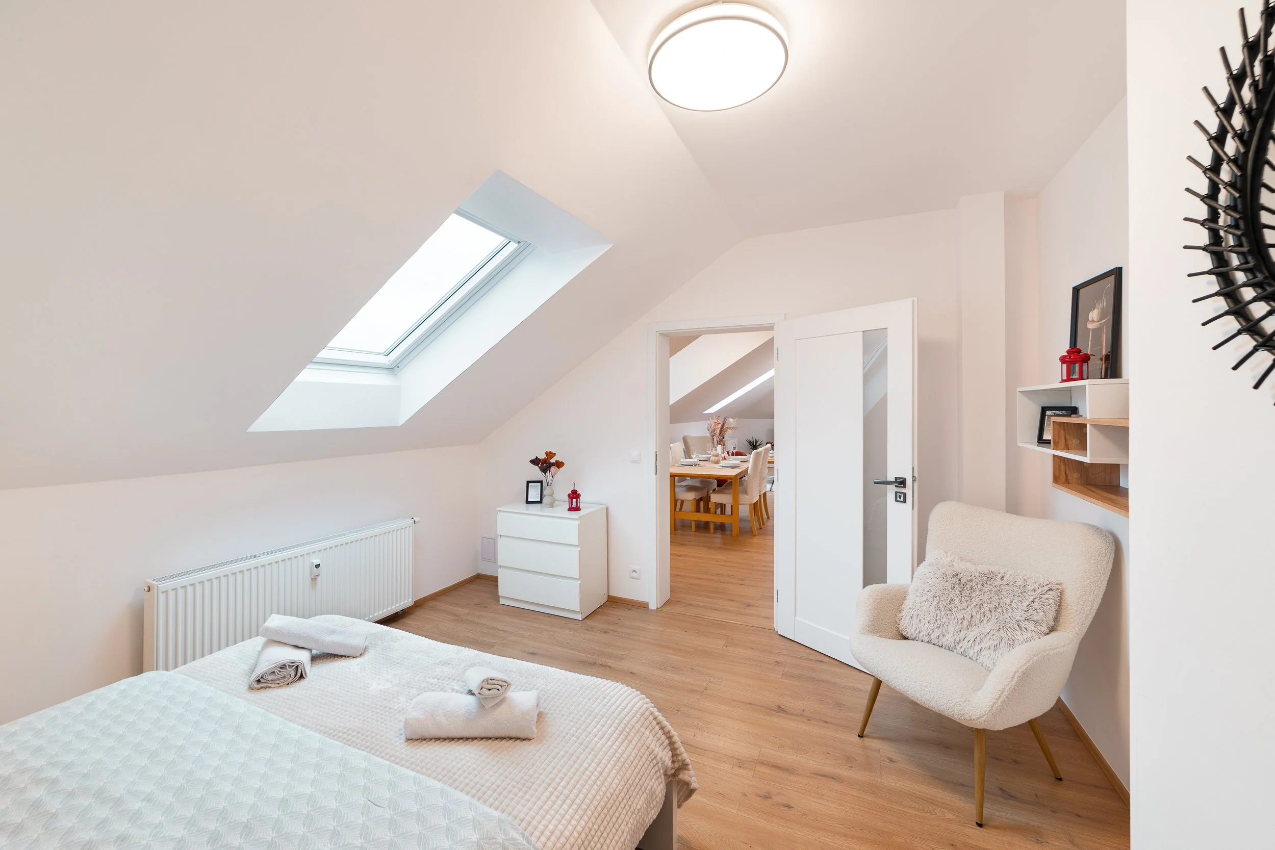 An attic bedroom with white walls and ceiling, skylight window, wooden flooring, a bed with white bedding and towels, a white dresser with decorative items, a beige armchair with a fluffy pillow, and a view into a dining area with a wooden table and 