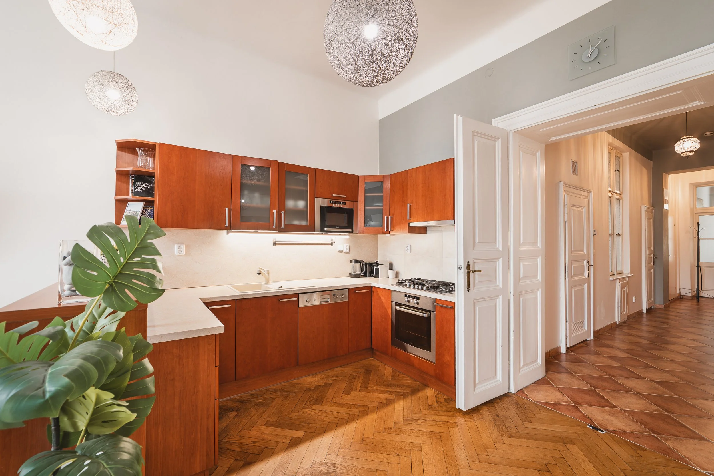 Modern kitchen with wooden cabinets, built-in oven and microwave, white countertops, green houseplant in the foreground, white walls, parquet wooden floor, and partially open white door leading to a hallway.