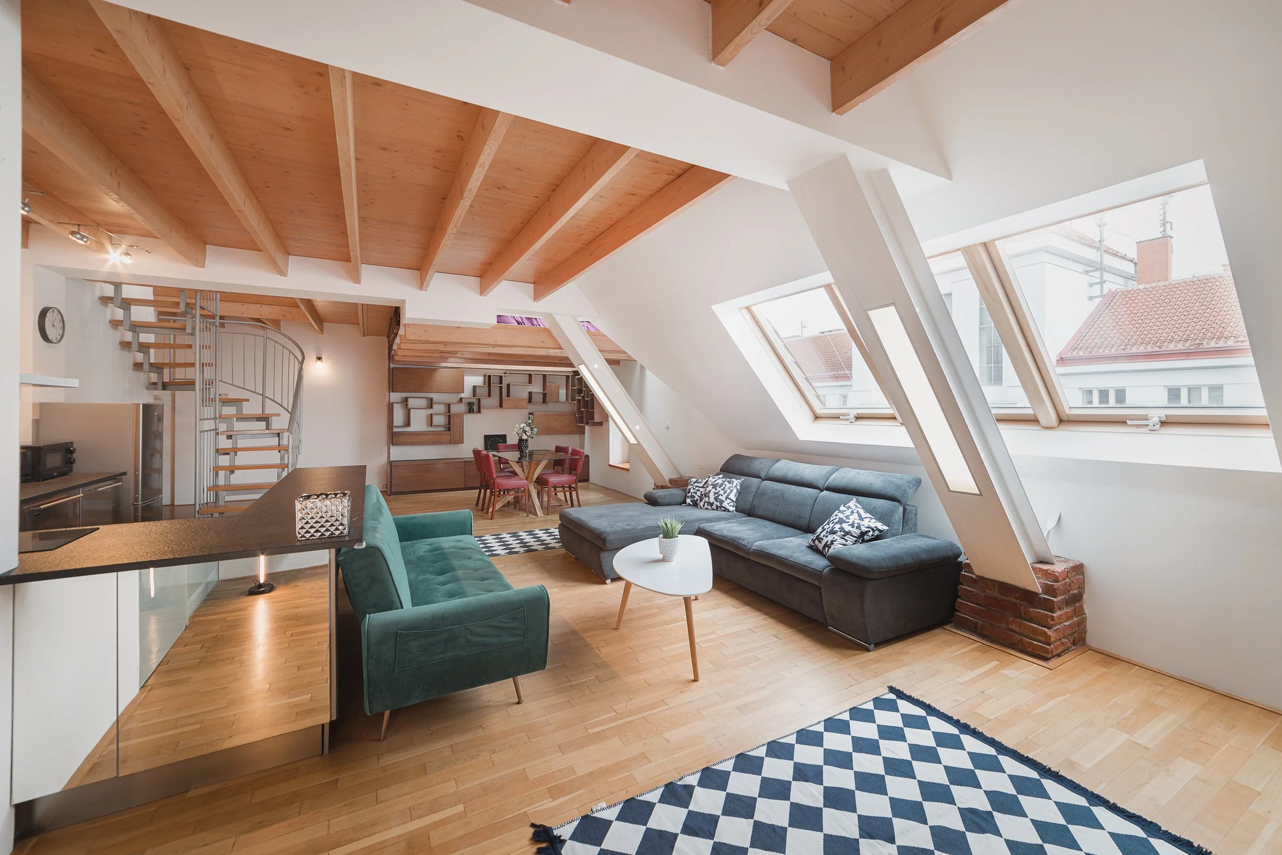 Living room with wooden ceiling beams, skylight windows, a gray sectional sofa, a green armchair, a round white coffee table, a black and white patterned rug, and a dining area with a round table and red chairs.