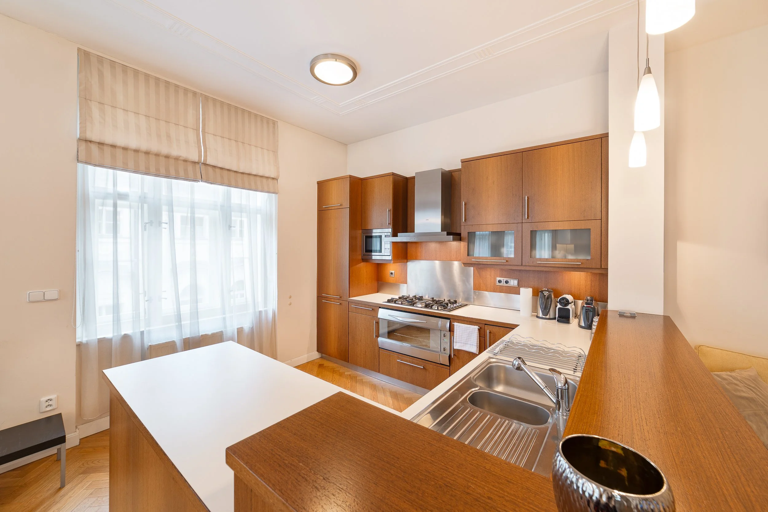 Modern kitchen with wooden cabinets, white countertop, stainless steel appliances, and a double sink, illuminated by ceiling and pendant lights, with a window covered by beige blinds and sheer curtains.