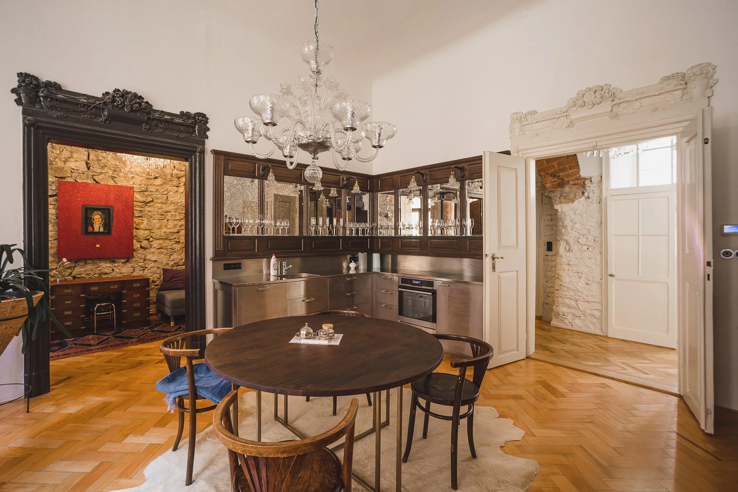 Interior of a modern kitchen with a dark wooden upper cabinet, stainless steel appliances, a chandelier, and a round dining table on a white rug.