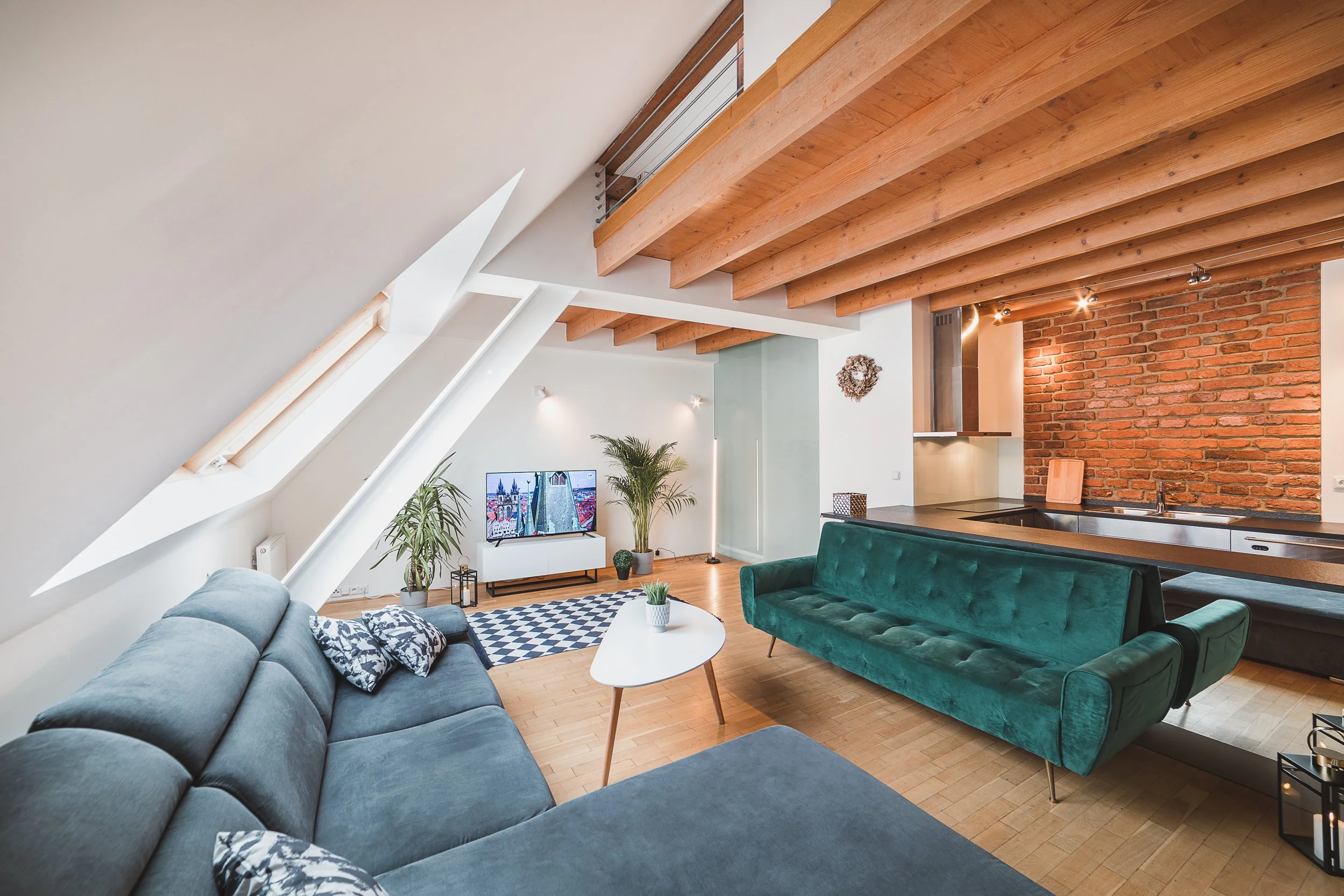 Living room with sloped ceiling, exposed wooden beams, brick wall, modern furniture, plants, and a television.