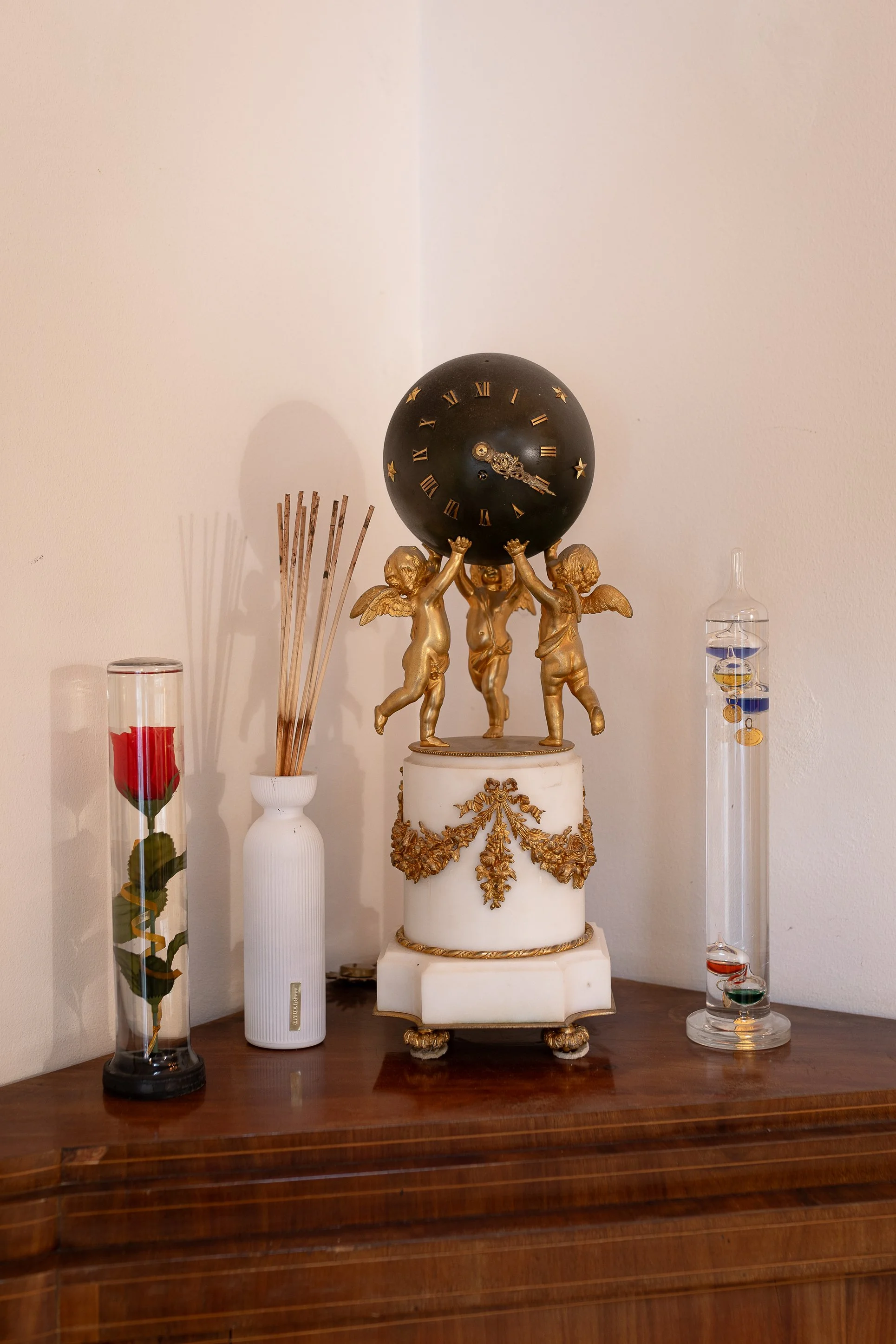 Decorative clock with gold cherubs holding a black ball clock with Roman numerals, placed on a wooden surface with vases and glass art pieces around it.