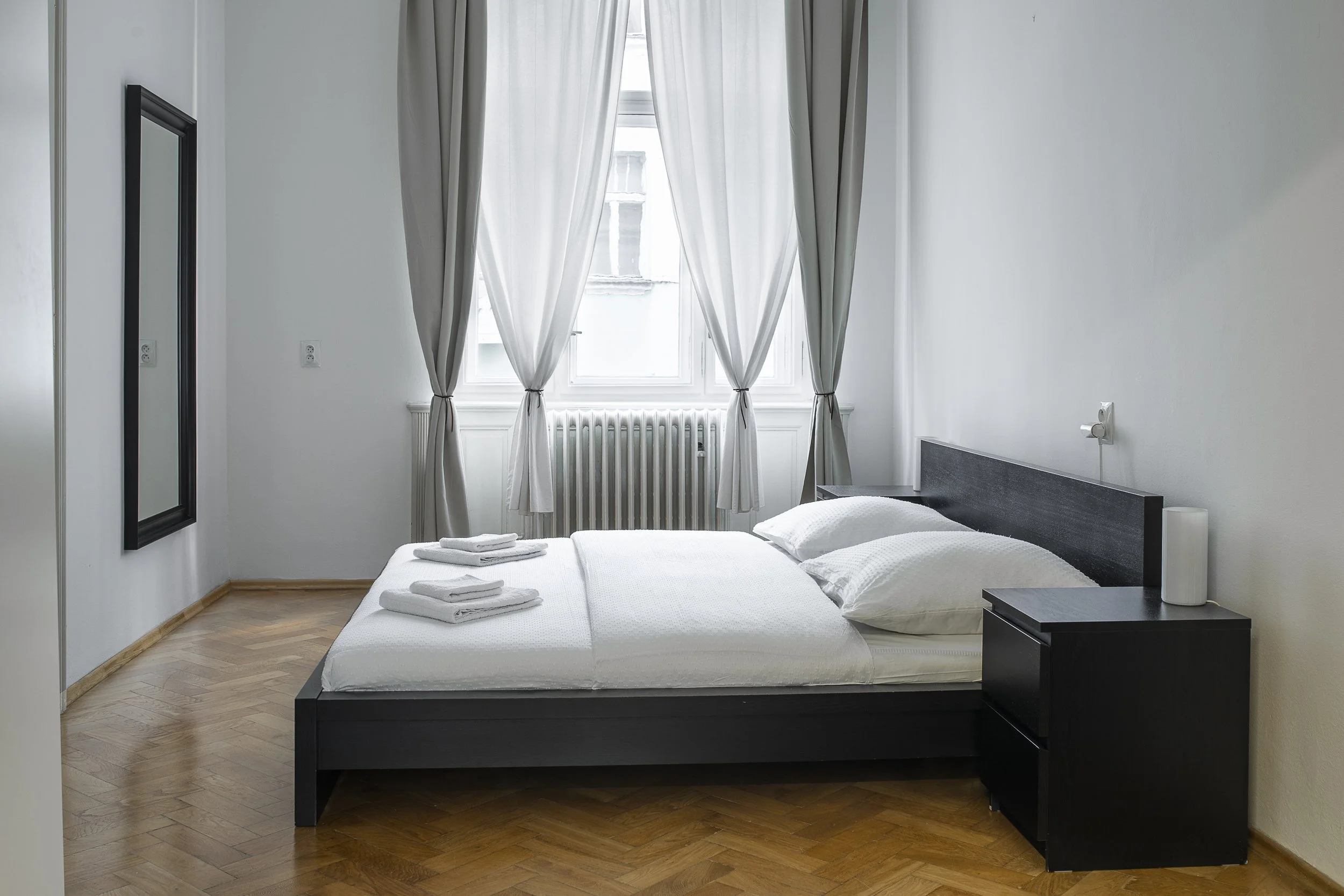 A minimalist bedroom with a large window covered by white curtains, a black bed frame with white bedding, neatly folded towels on the bed, a black nightstand with a white lamp, and light wooden floors.