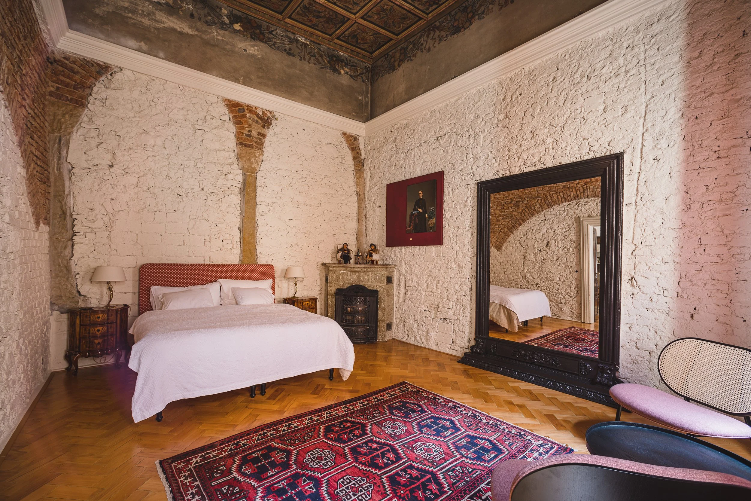 A bedroom with exposed brick walls, a large mirror, and a bed with a red and white headboard. There are two nightstands with lamps, a portrait hanging on the wall, a decorative fireplace, and a Persian rug on the wooden floor.