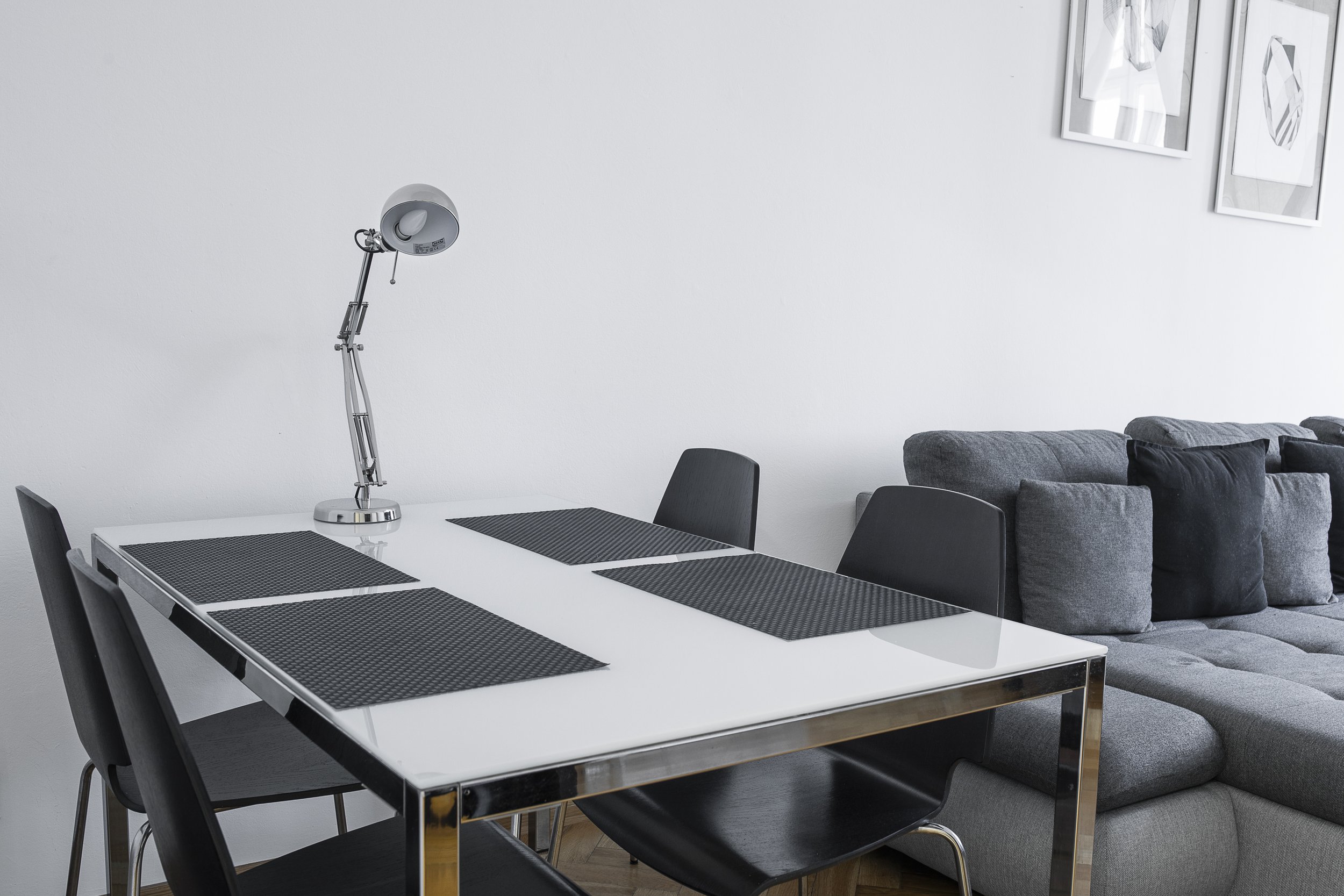 A modern dining room with a white table, black chairs, and black placemats. A sleek, adjustable silver desk lamp is on the table. Behind the table, there is a gray sofa with black pillows. The wall behind features framed geometric artwork.