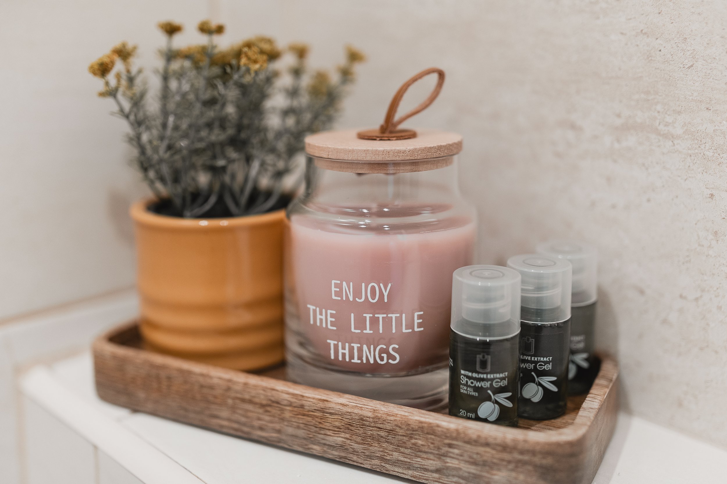 A wooden tray holding a pink candle with a wooden lid, small spray bottles of olive extract shower gel, and a potted plant with dried flowers.