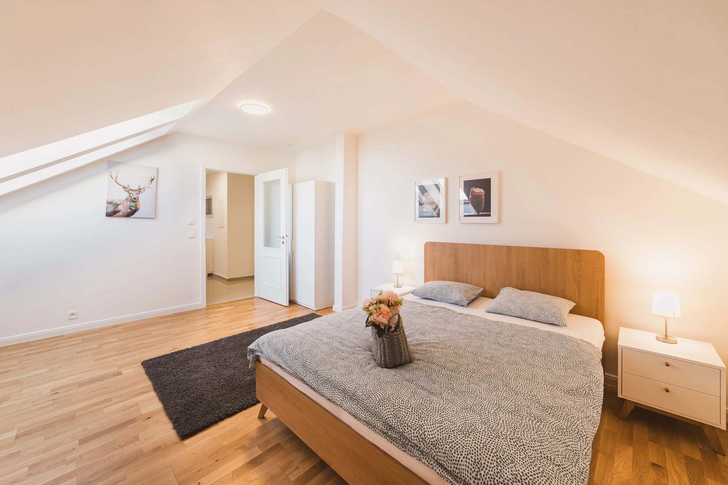 A bedroom with a wooden bed frame, patterned bedding, and a vase of flowers on a wicker basket. There are three framed pictures on the wall, a nightstand with a lamp, and a dark rug on the hardwood floor. The room has a sloped ceiling with skylights 