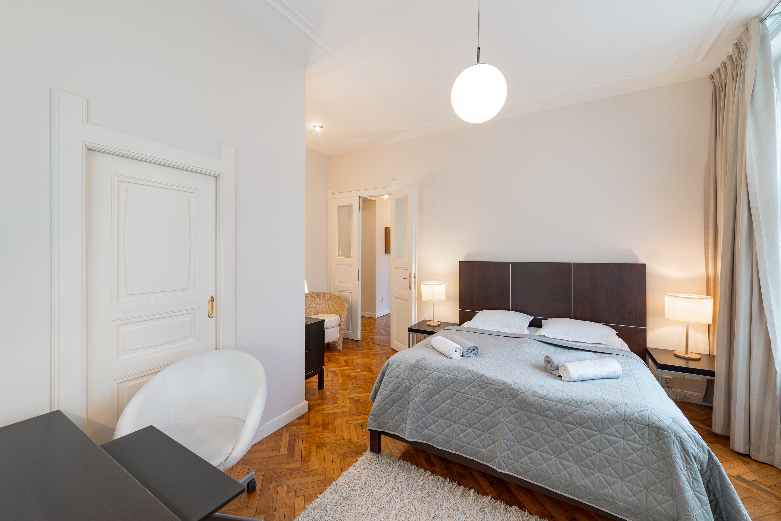 A bedroom with a bed, nightstands, lamps, desk, white chair, and curtains.