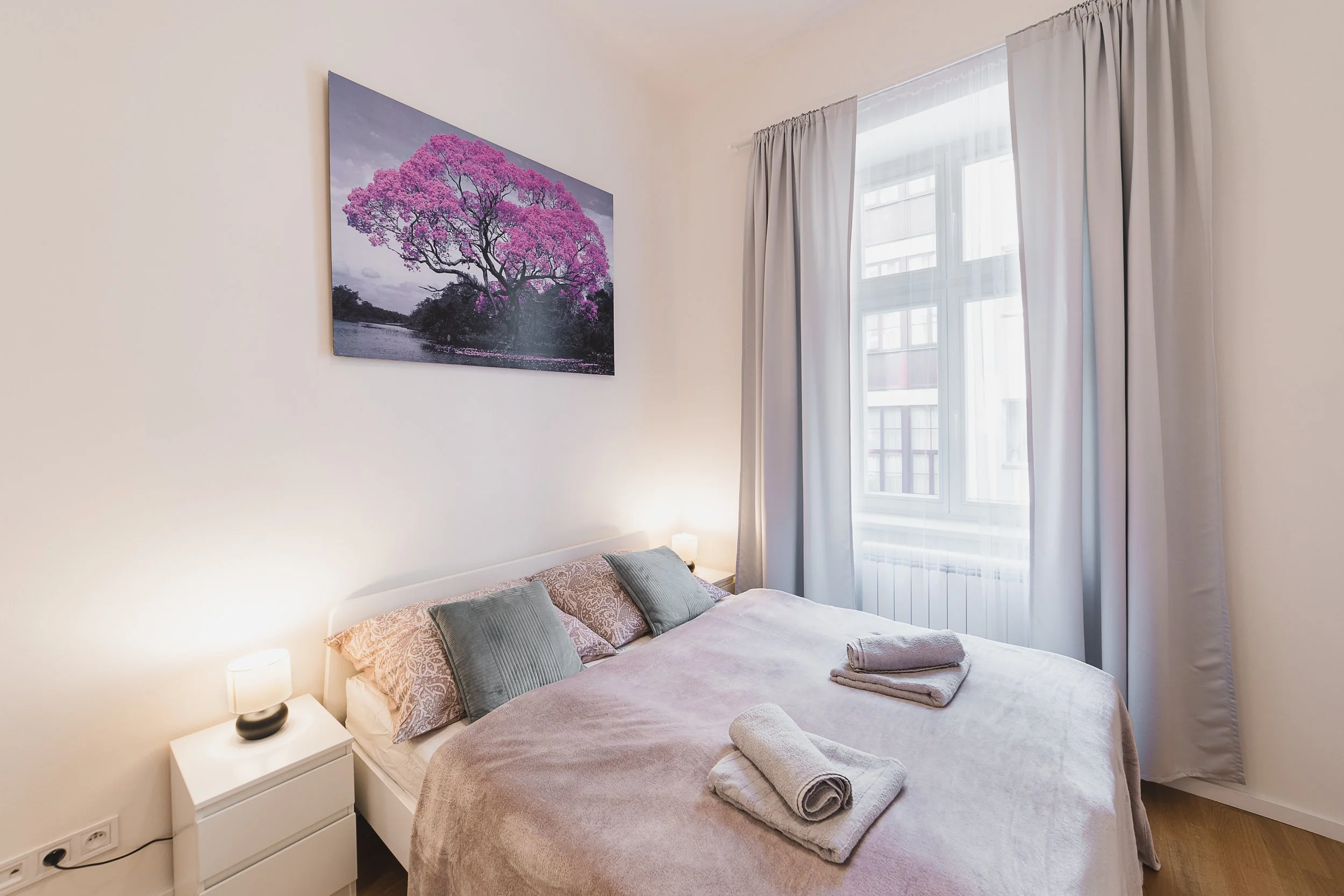 A neatly made bed with a light pink bedspread, two rolled towels, and cushions, next to a white nightstand with a small lamp, in a bright room with a large window and white curtains, and a picture of a pink flowering tree hanging on the wall.