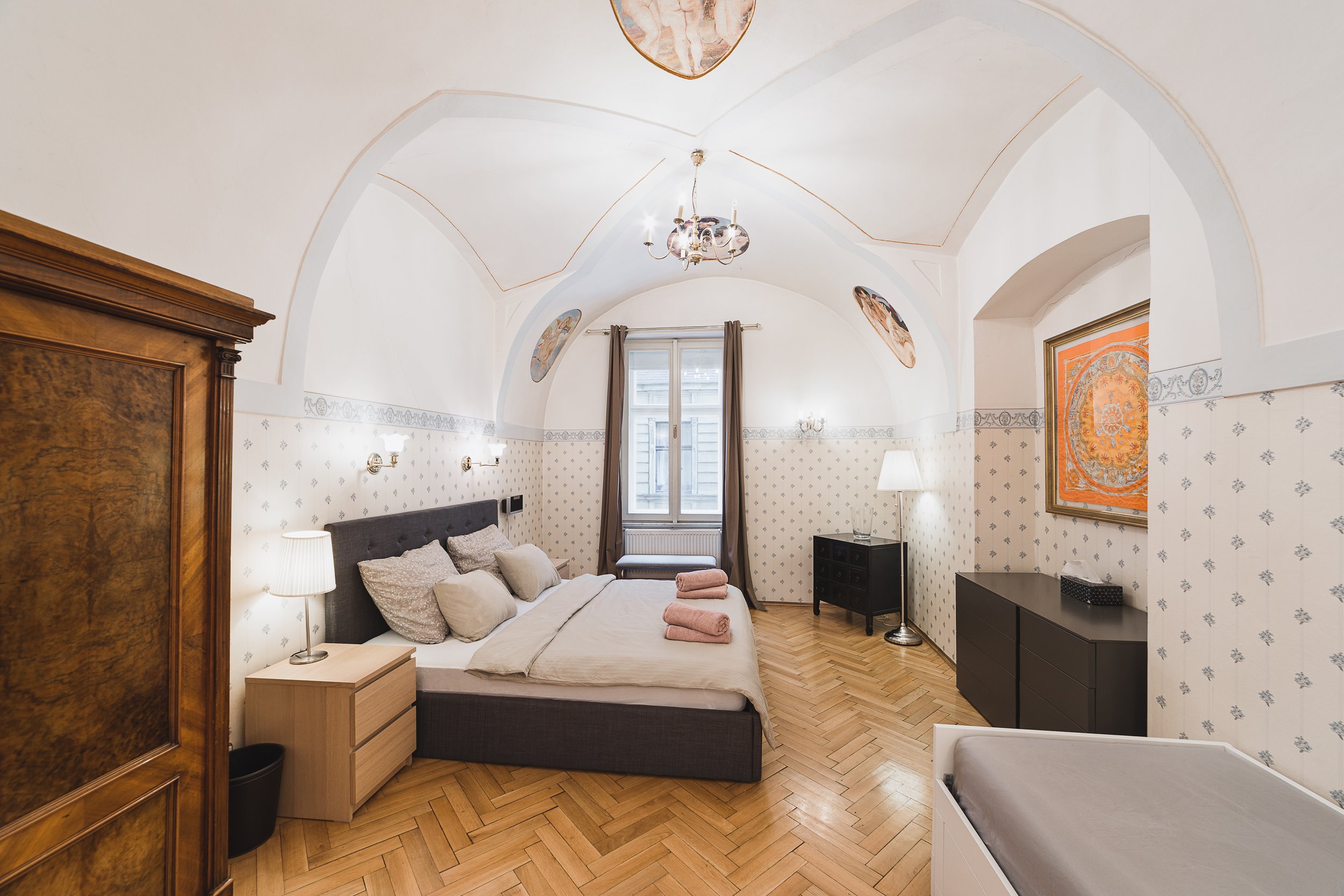 A cozy bedroom with a high arched ceiling, a large bed with beige and gray pillows, a wooden nightstand with a lamp, and a window with curtains. The room has decorative wall art, a chandelier, and a black dresser.