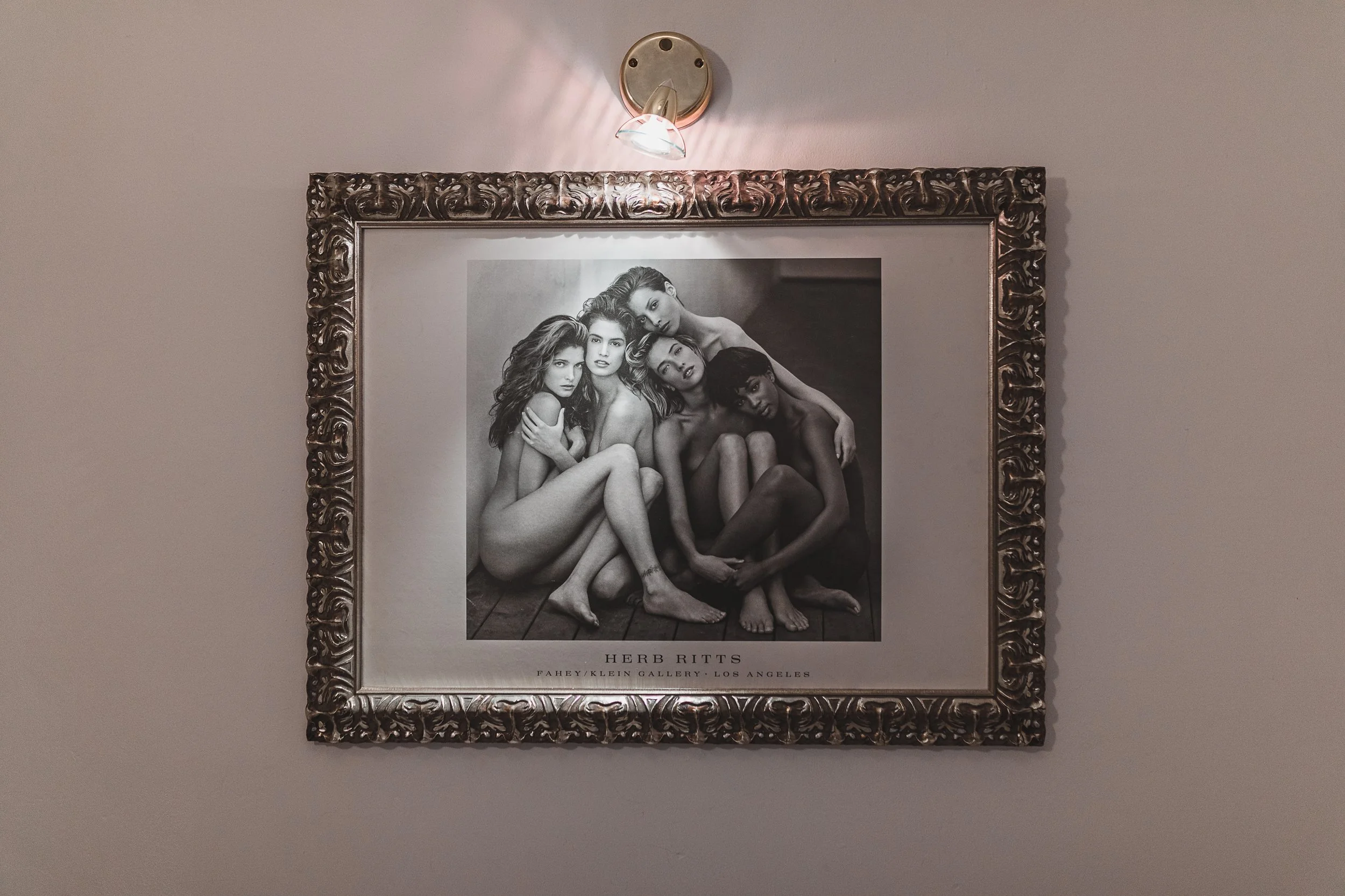 Black and white photograph of six women sitting close together with arms around each other. They are nude with legs crossed or bent, and their hair varies in length and style. The photo is framed in an ornate, decorative frame and is labeled with the