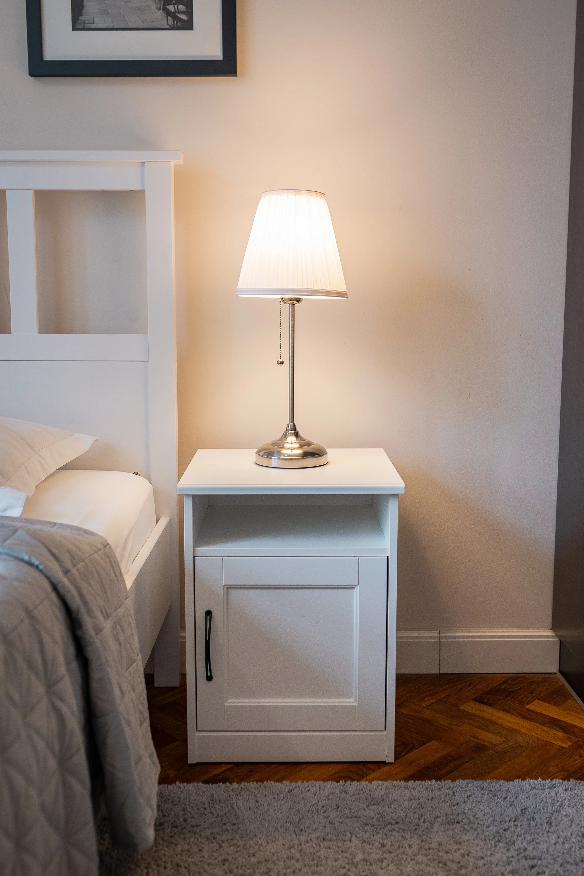 White nightstand with a lamp on top, situated next to a bed with beige and gray bedding, in a room with beige walls, hardwood floor, and a framed picture on the wall.