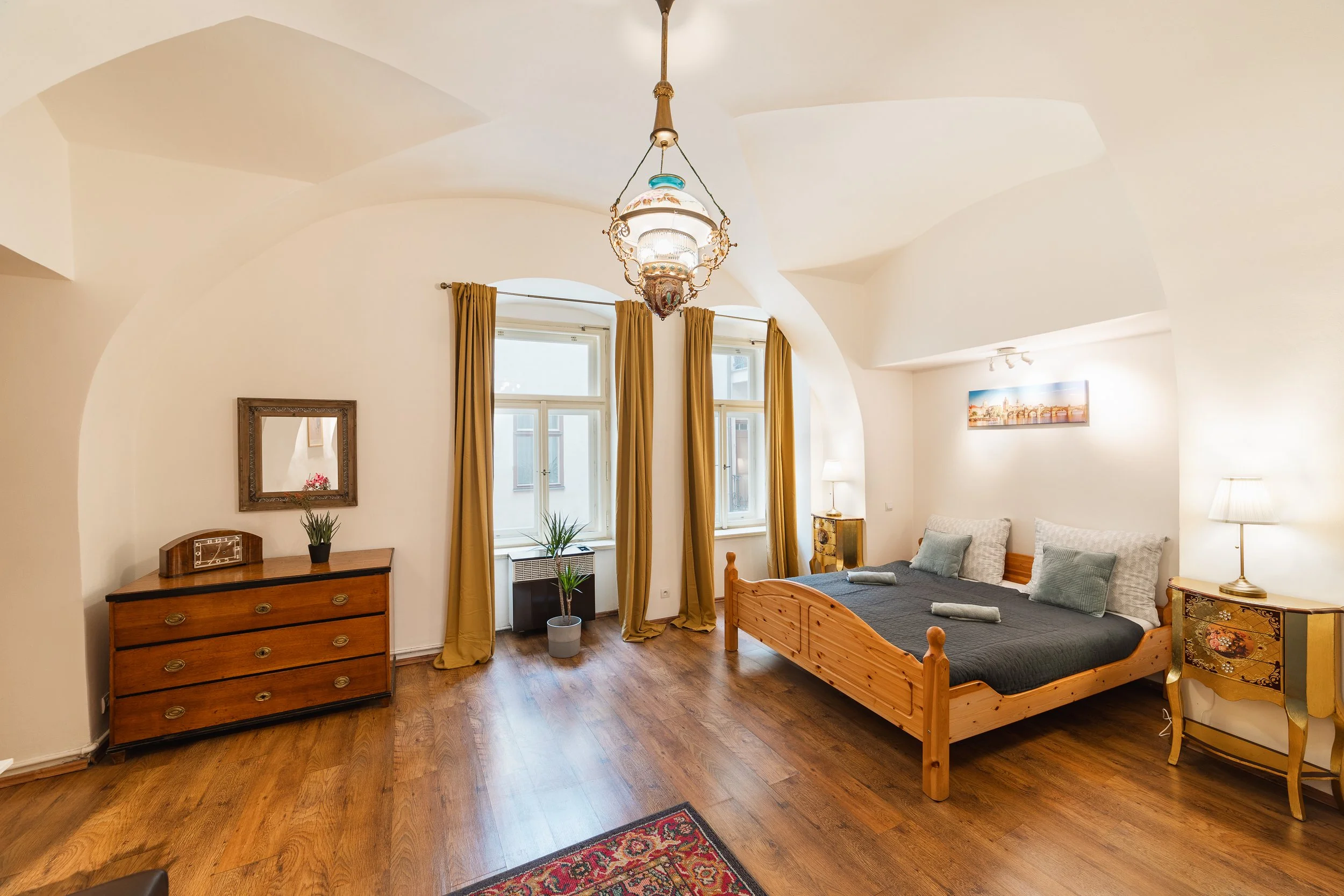 A bedroom with wooden floors, a wooden double bed with bedding, a wooden dresser, framed artwork, plants, a hanging light fixture, large windows with yellow curtains, and a small decorative table.