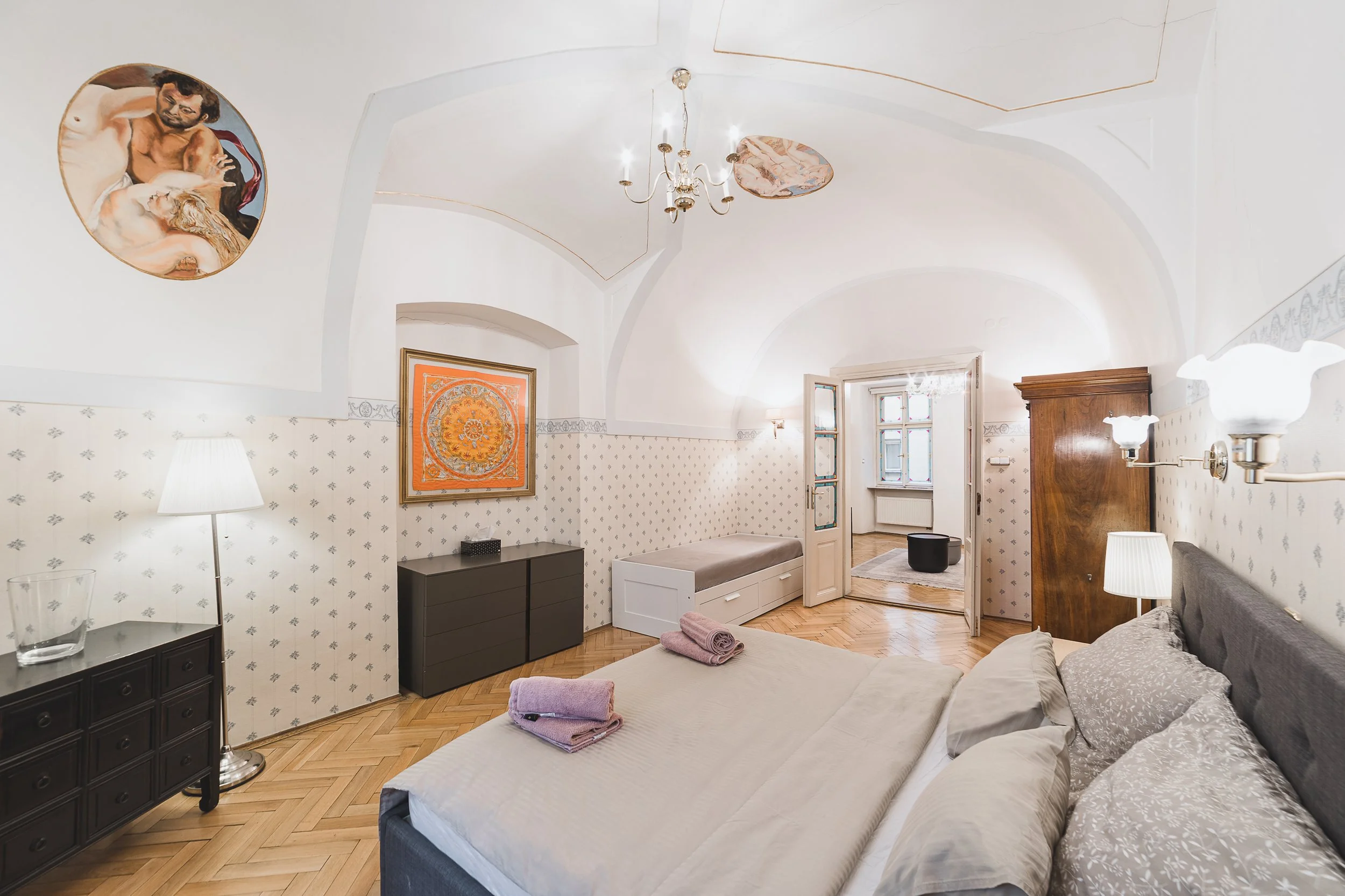 A bright, spacious bedroom with white walls and ornate ceiling design. The bed has beige linens, multiple pillows, and folded towels. There are two lamps, a black side table, and artwork on the walls, including a framed picture and two circular paint
