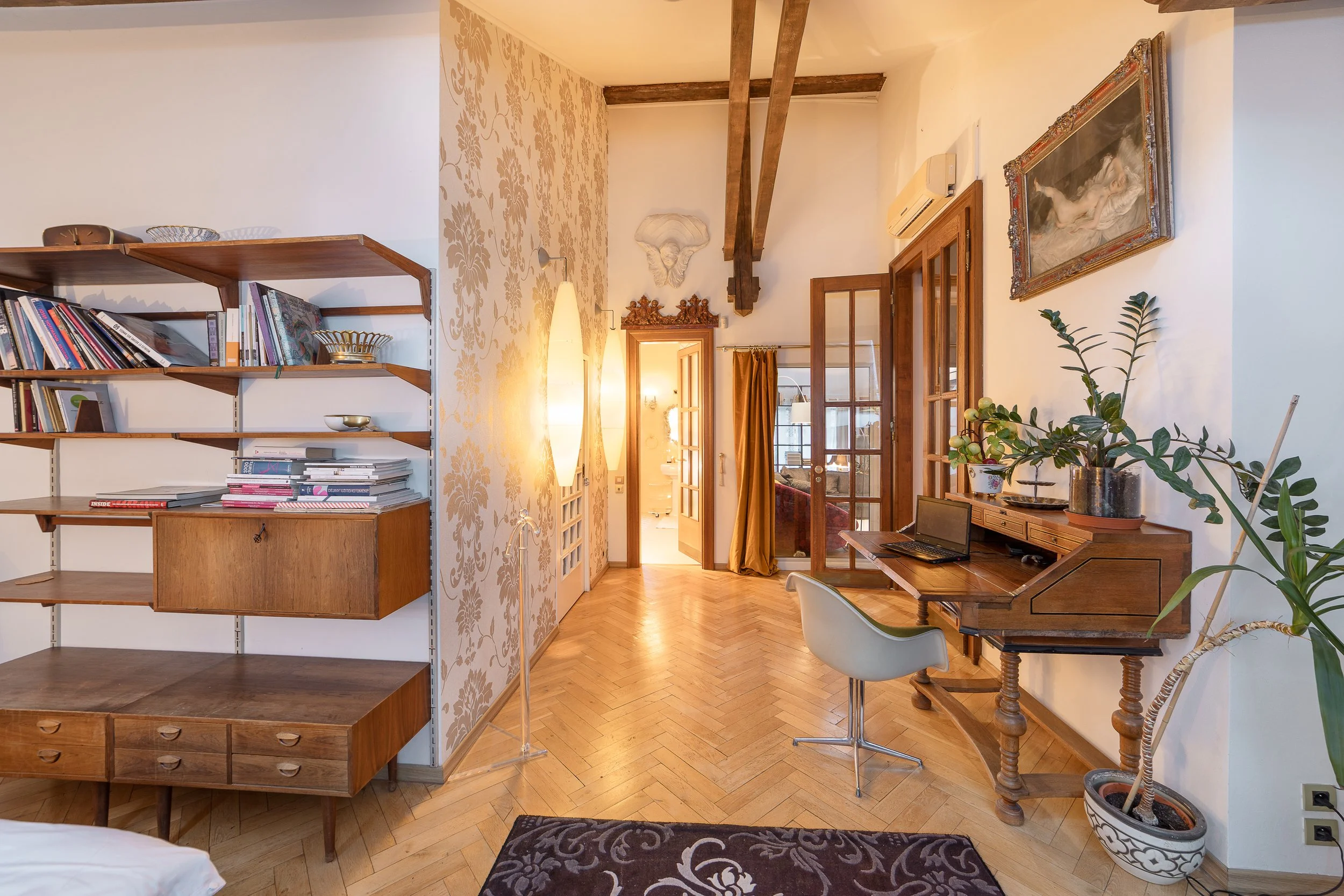 Interior view of a living space with wooden flooring, a bookshelf, a wooden desk with a computer, and a potted plant, decorated with artwork on the walls and warm lighting.